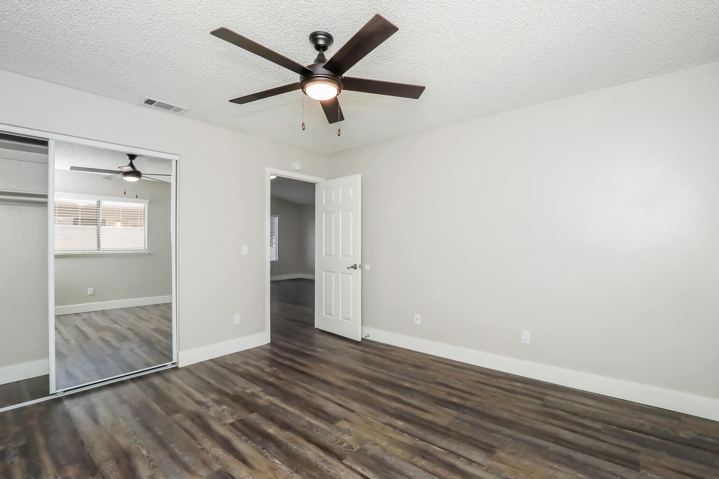 A spacious, unfurnished room featuring light-colored walls, a ceiling fan, and large mirror closet doors. The floor is dark hardwood, and there is an open door leading to another area. Bright natural light floods the space, enhancing its inviting atmosphere.