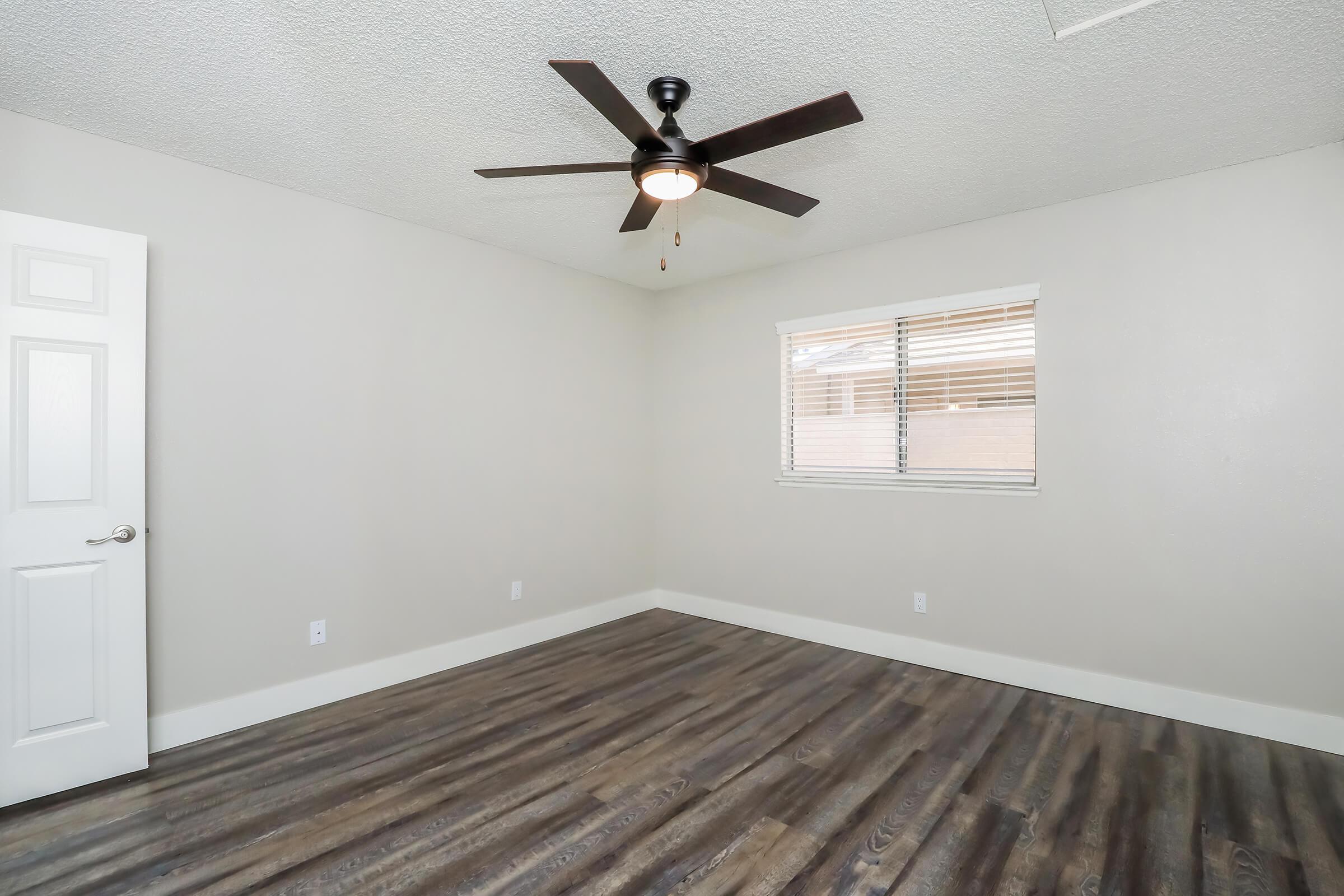 A bright, empty room with light-colored walls and a ceiling fan. There is a window with blinds allowing natural light in, and modern laminate flooring. A white door is visible on the left side of the image, enhancing the spacious feeling of the room.