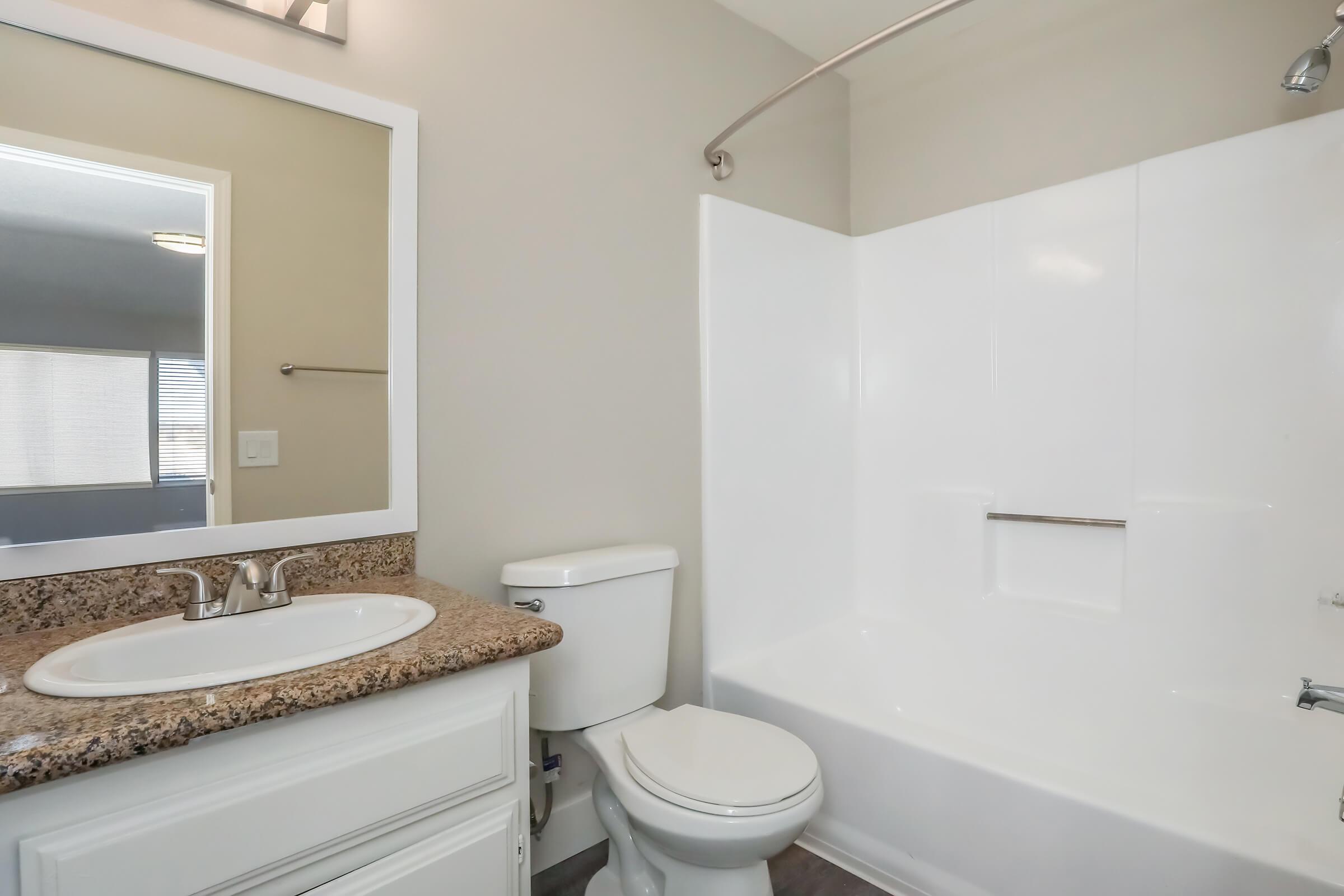 Bright and clean bathroom featuring a white bathtub with a shower, a toilet, and a sink with a granite countertop. There is a large mirror above the sink and light-colored walls, creating a spacious and modern feel. Natural light comes through a nearby window.