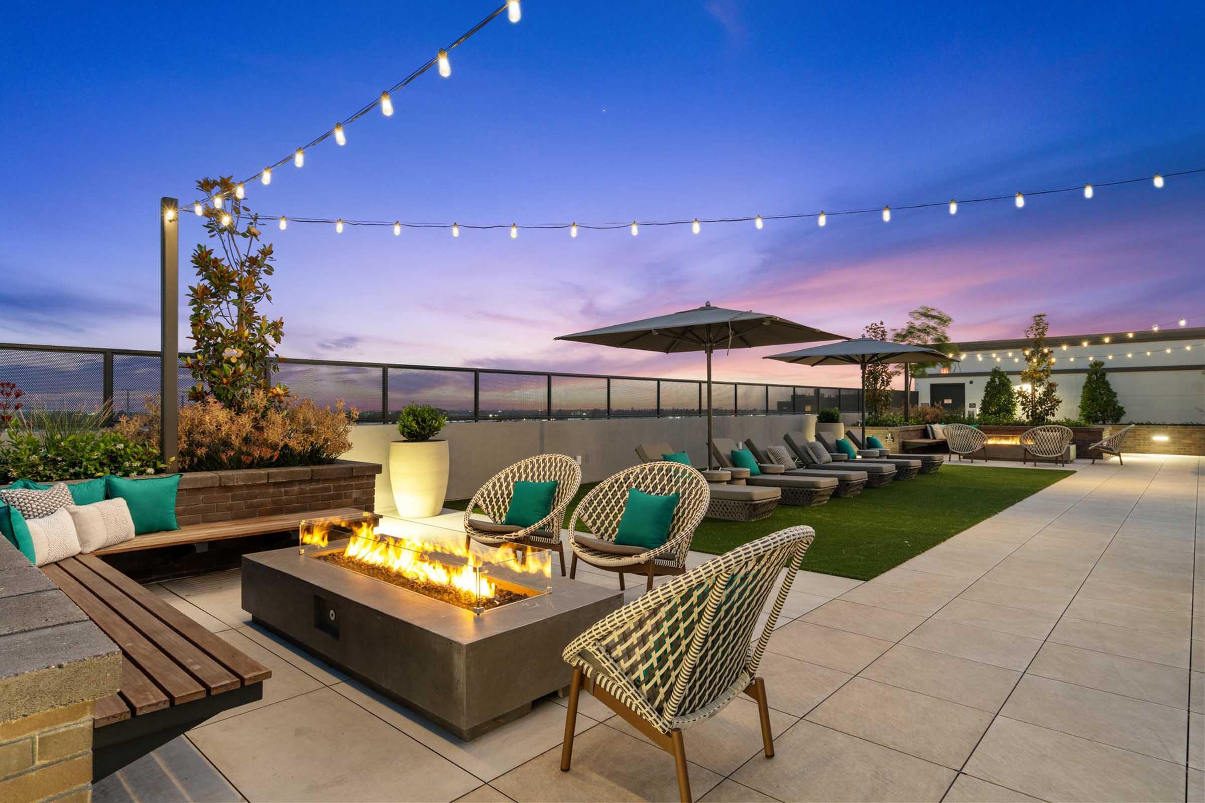 A modern rooftop lounge featuring a fire pit with flames, surrounded by stylish seating and cozy pillows. The area is adorned with string lights and green plants, set against a vibrant sunset sky, creating a warm and inviting atmosphere for relaxation and socializing.