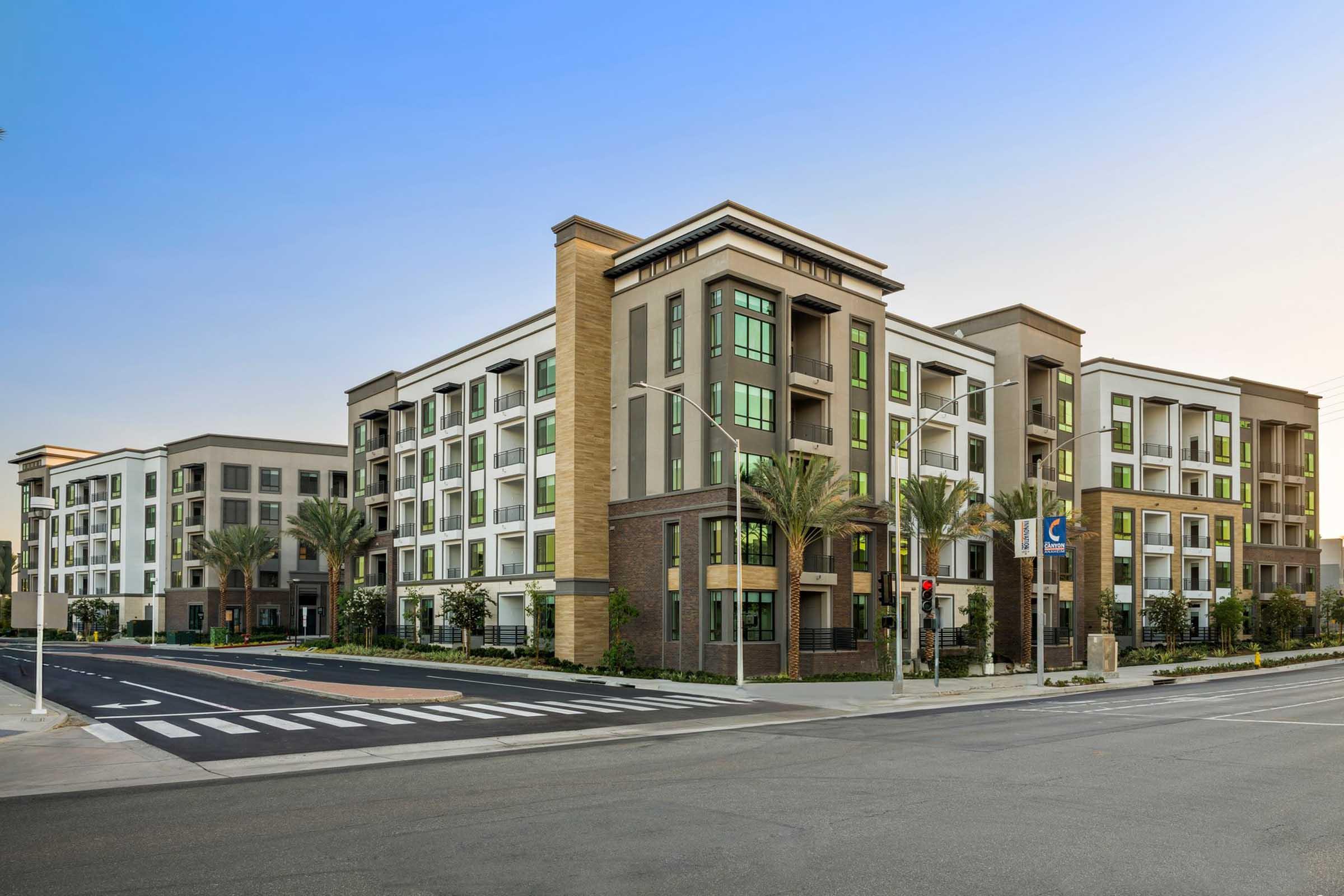 Modern multifamily residential buildings in a suburban setting, featuring contemporary architecture with a mix of brick and stucco. The complex has several floors, large windows, and palm trees lining the walkways. A quiet street with traffic lights and crosswalks is visible in the foreground, under a clear blue sky.