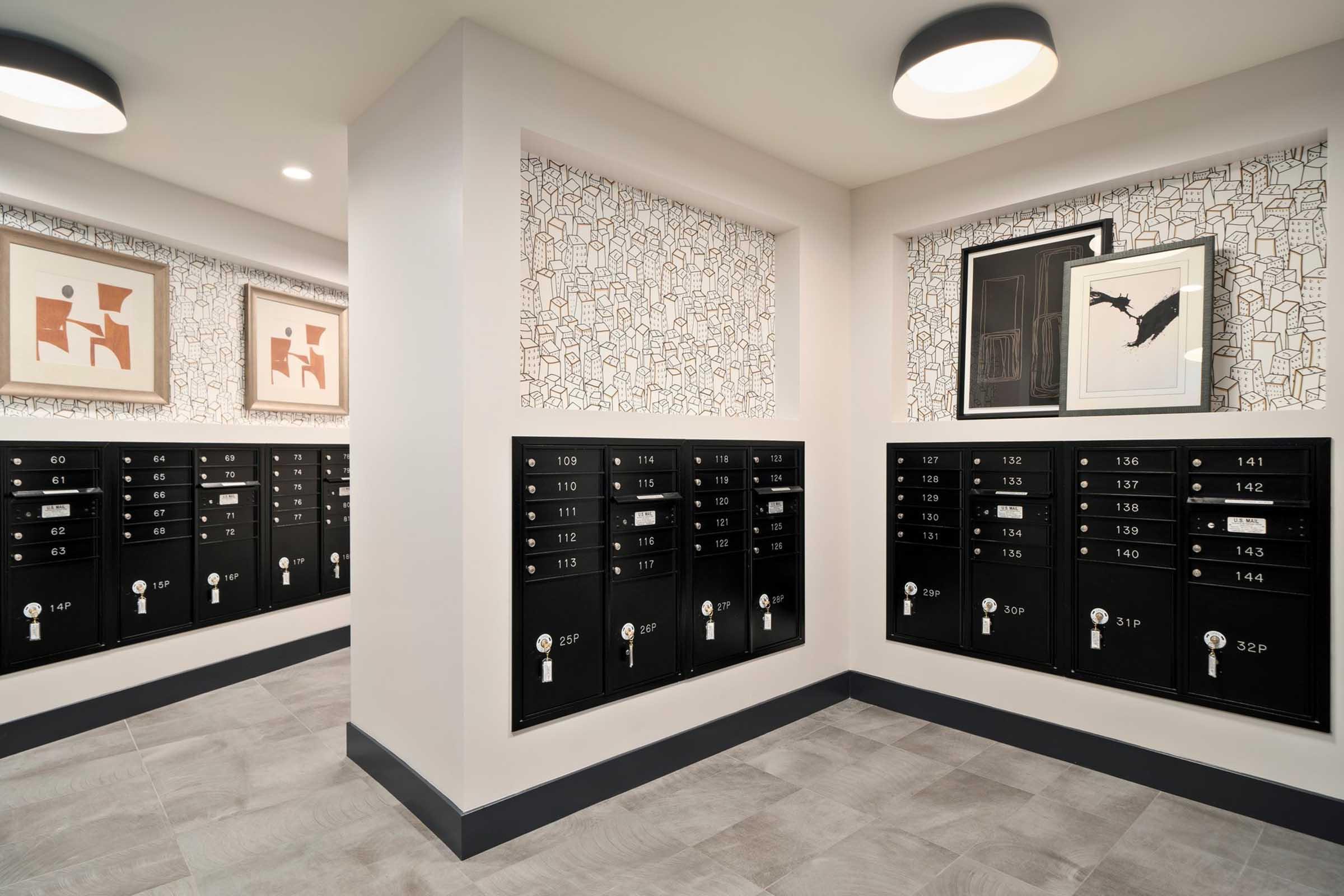 Interior view of a mailbox area featuring several black postal boxes mounted on the wall. The space is well-lit with round ceiling lights, and there are decorative framed artworks on the walls, all set against a textured, patterned wallpaper. The flooring is tiled in light gray.