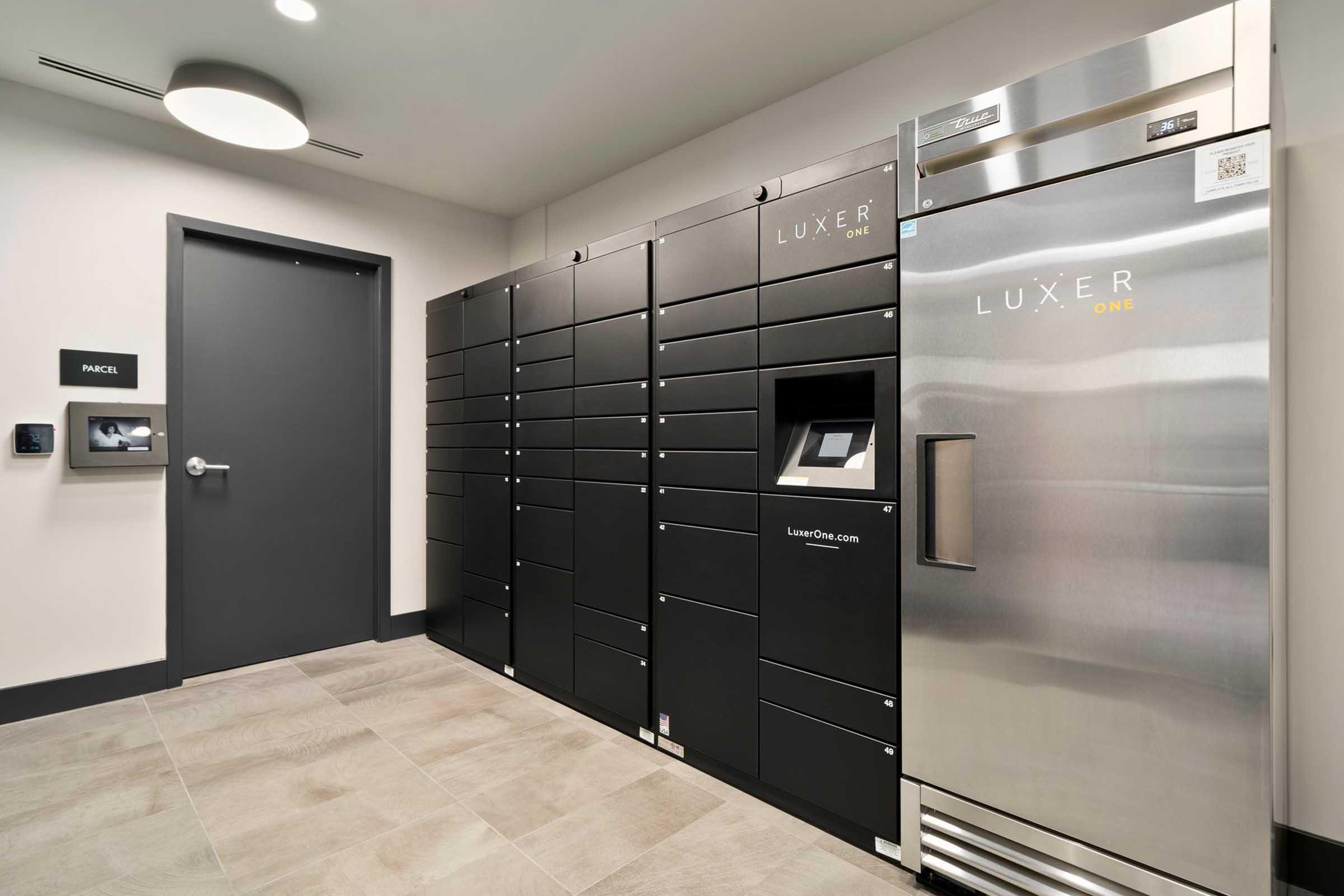 A modern delivery locker system with multiple black lockers and a digital interface, alongside a metallic refrigerator. The space features neutral-colored walls and flooring, with a door labeled "RANCH" and a wall-mounted control panel nearby. Ideal for package delivery and secure storage.
