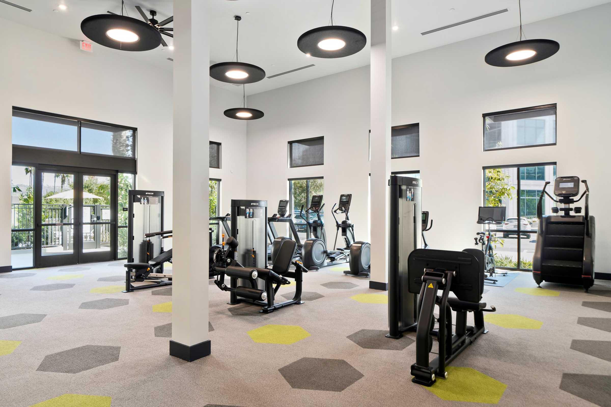 A modern gym interior featuring various fitness equipment, including treadmills, elliptical machines, and weight training machines, set against a bright, airy space with large windows. The floor is covered with gray carpeting and yellow hexagonal accents, and there are ceiling fans and pendant lights for illumination.