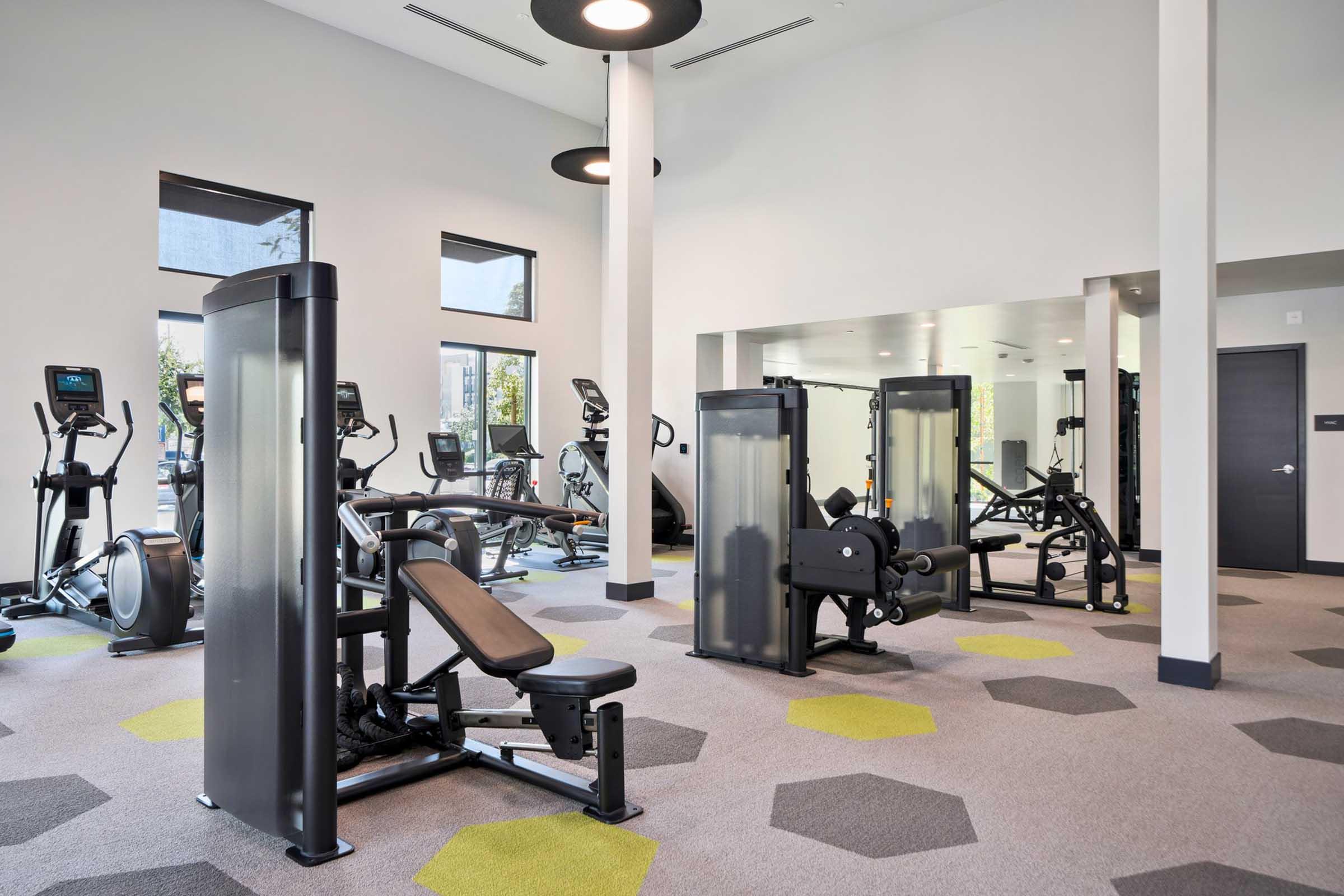 A modern gym interior featuring a variety of fitness equipment, including elliptical machines, exercise bikes, and weightlifting machines. The space is well-lit with large windows and has a clean, contemporary design, with hexagonal-patterned flooring in shades of gray and green.