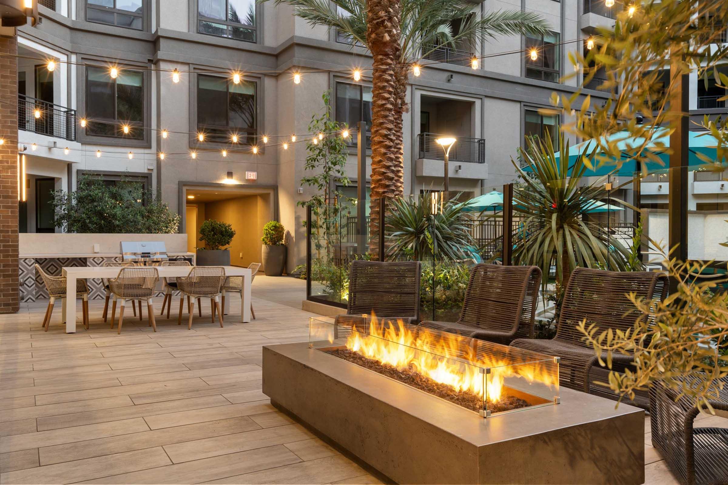 A modern outdoor lounge area featuring a fire pit with flames, surrounded by stylish seating. Lush plants and palm trees are present, along with string lights overhead. In the background, there's a dining table and chairs, creating a cozy and inviting atmosphere. The setting is designed for relaxation and socializing.