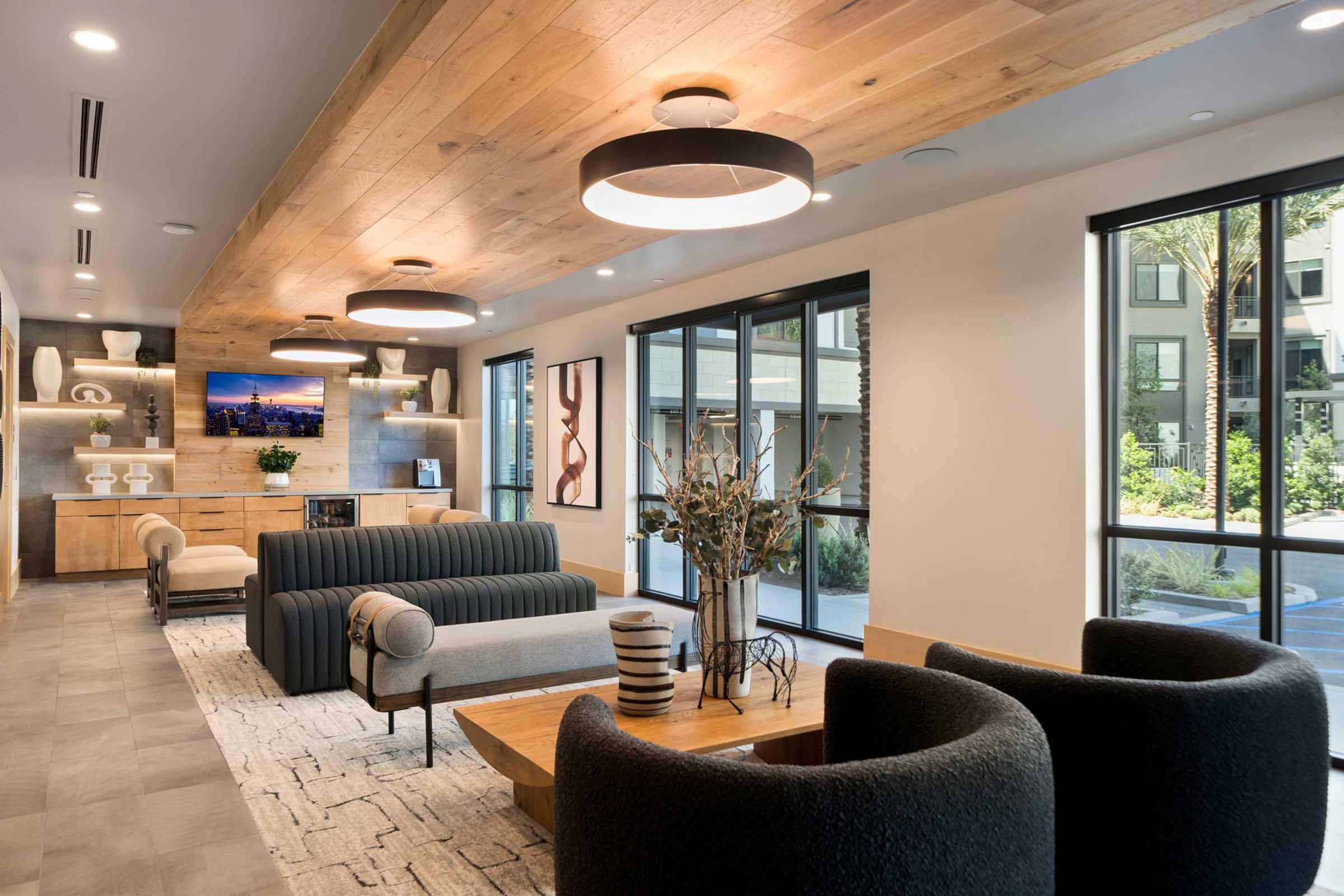 A modern, stylish living space featuring a striped sofa, two black armchairs, and a wooden coffee table. The room has wooden beam ceilings, large windows overlooking an outdoor space, and decorative elements including a vase with flowers and contemporary artwork on the walls.