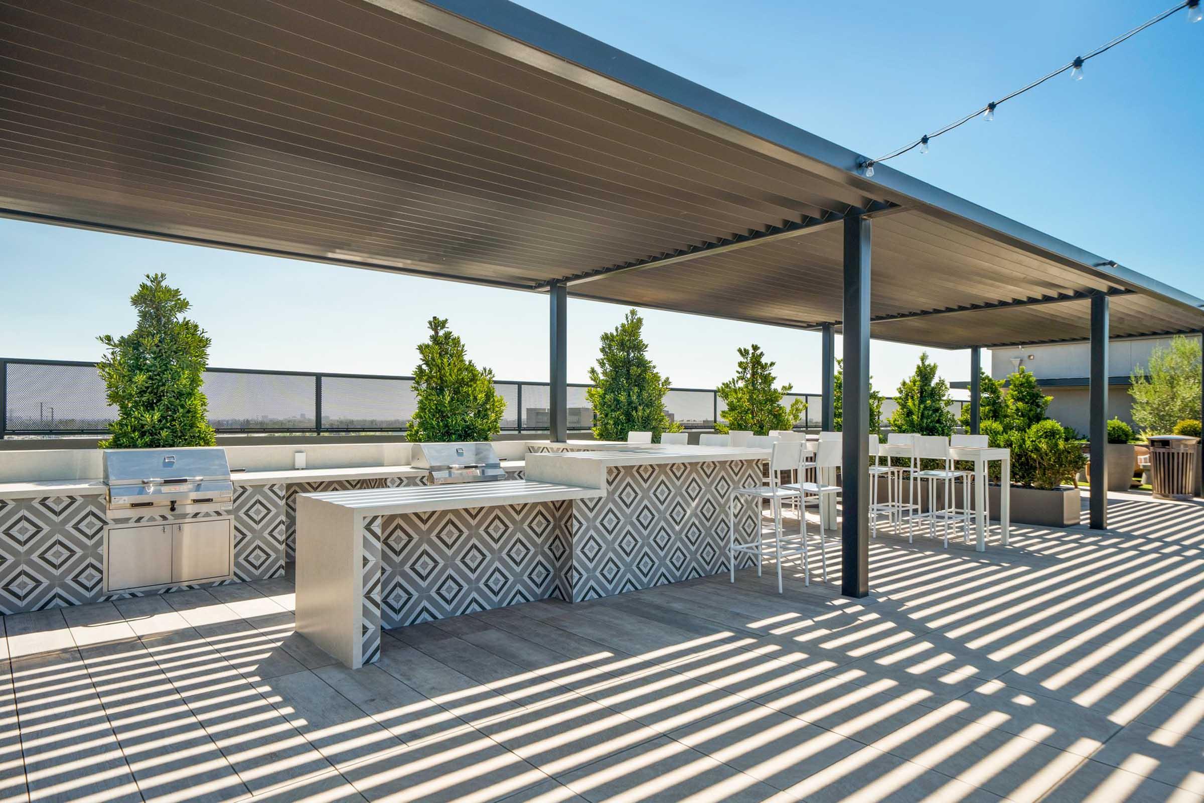 A modern rooftop terrace featuring a stylish outdoor kitchen with a grill and bar seating. The area is decorated with geometric patterned tiles and surrounded by greenery, under a shaded pergola. Sunlight creates shadows on the tiled floor, enhancing the inviting atmosphere.
