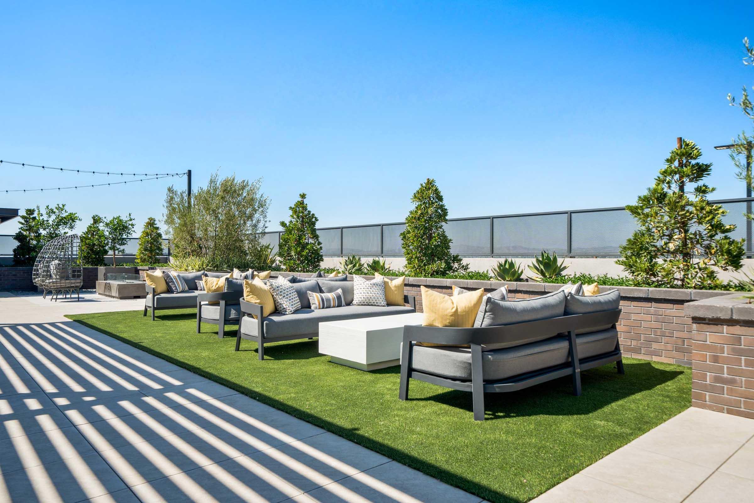 A modern rooftop lounge area featuring several gray sofas with yellow accent pillows, arranged on artificial grass. The space is surrounded by green plants and a low brick wall, under a bright blue sky. Soft shadows from overhead structures create patterns on the ground.