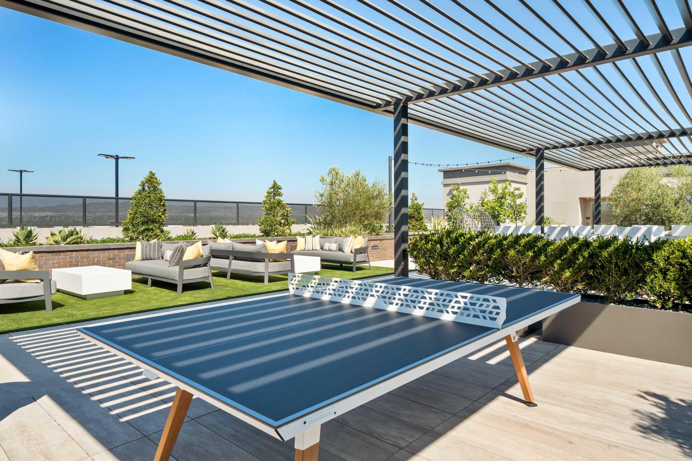 A rooftop terrace featuring a ping pong table, modern seating areas with cushions, green plants, and trees under a pergola. The setting is bright and sunny, with a clear blue sky in the background, creating a relaxing atmosphere for socializing and leisure activities.