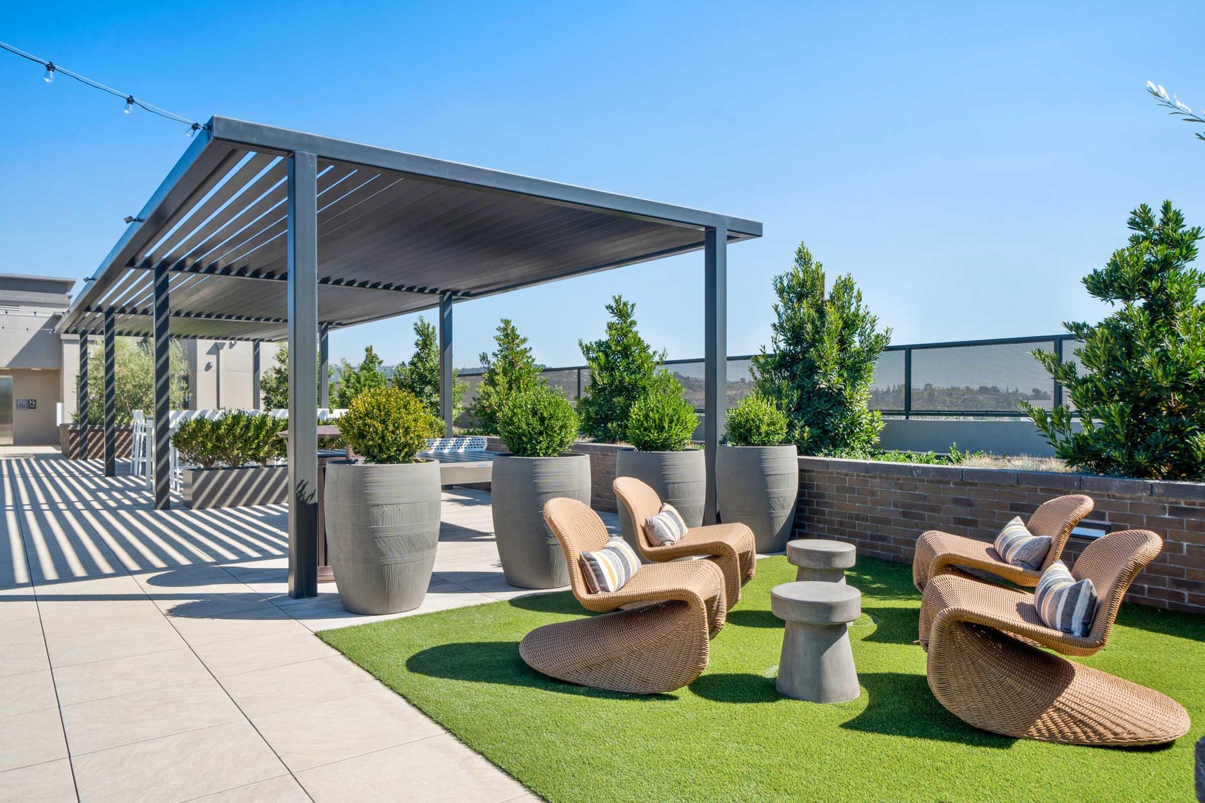 A rooftop terrace featuring modern wicker chairs with cushions, a small stone table, and large potted plants under a shaded pergola. The area is designed for outdoor relaxation, with green grass flooring and clear blue sky in the background.