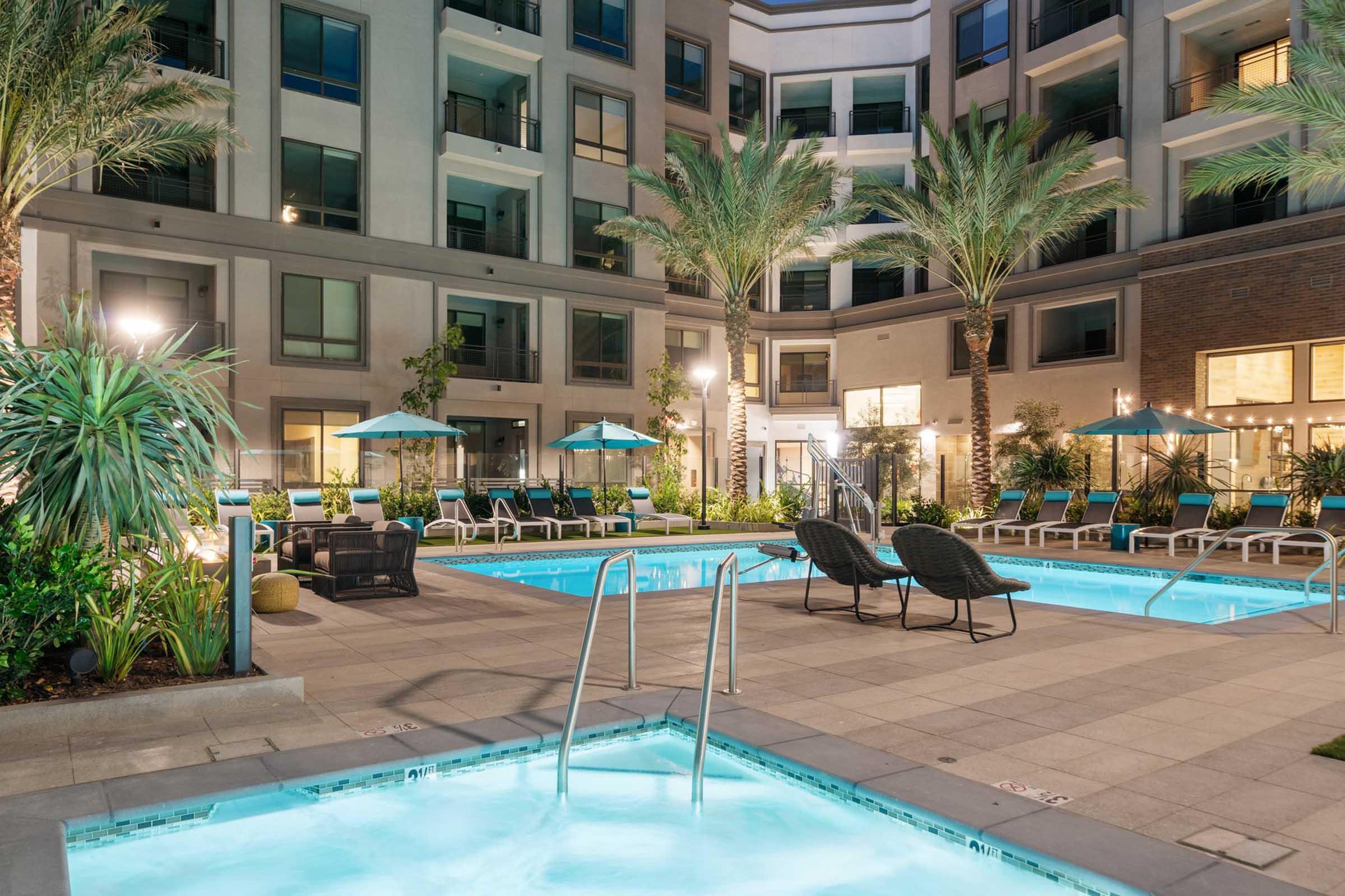 A modern apartment complex courtyard featuring two pools, surrounded by palm trees and lounge chairs. Soft lighting enhances the evening ambiance, while umbrellas provide shade. The area is designed for relaxation and socializing, with well-maintained landscaping.