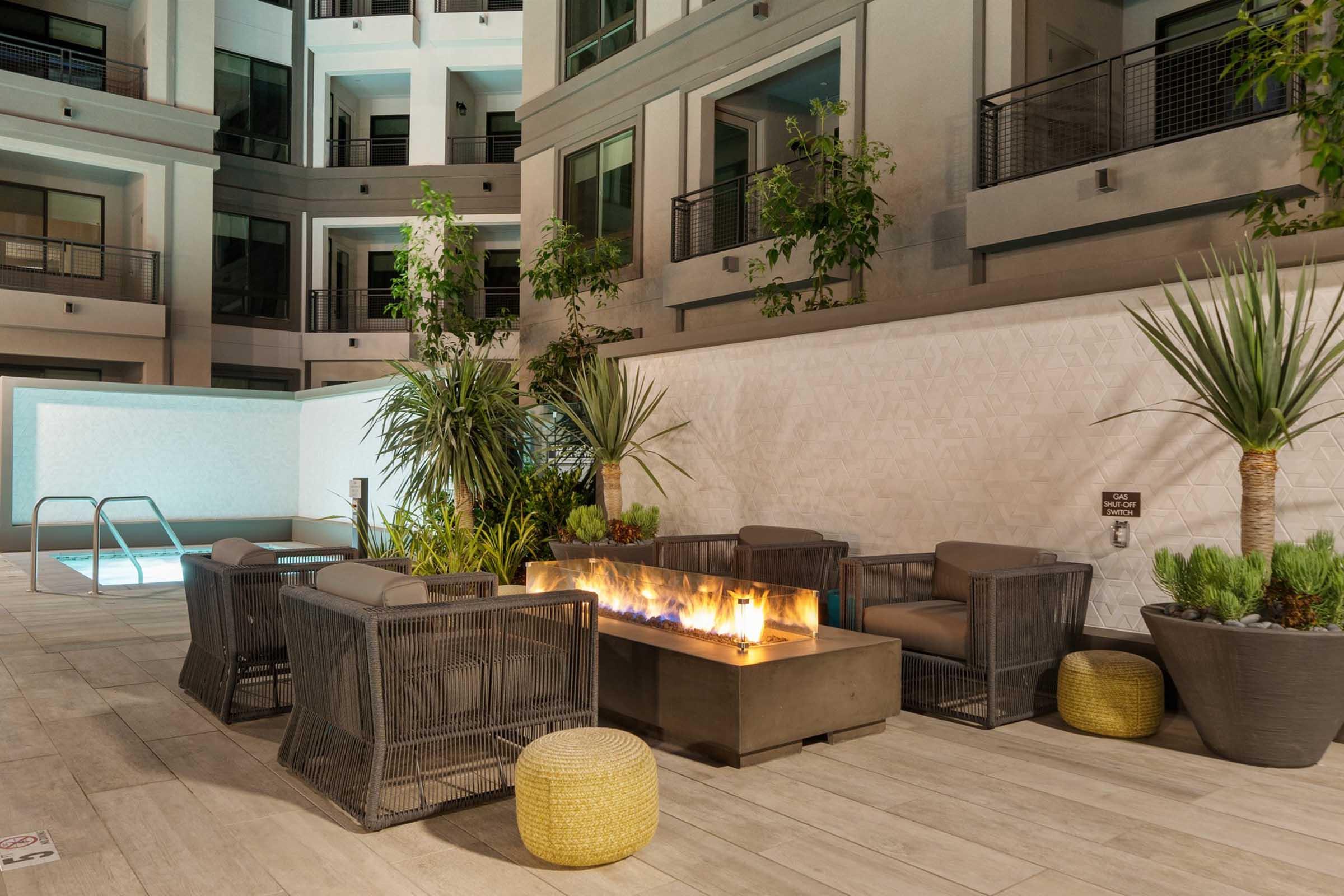 A modern outdoor patio featuring a cozy seating area with gray chairs around a fire pit, surrounded by lush greenery. In the background, a pool is visible along with contemporary apartment building facades, creating a stylish and inviting atmosphere for relaxation.