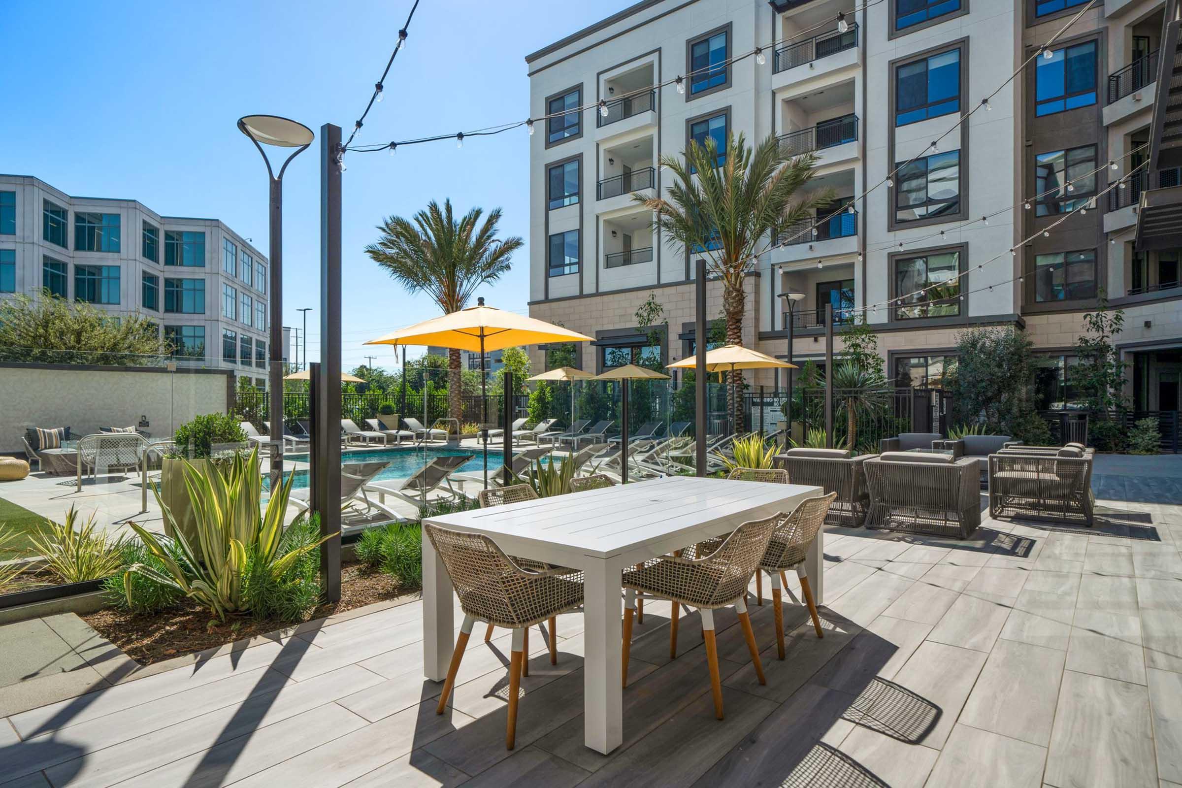A sunny outdoor pool area at an apartment complex featuring lounge chairs, palm trees, and large umbrellas. A white dining table with woven chairs is set near the pool, surrounded by greenery and modern architecture. Bright blue skies and well-maintained landscaping create a welcoming atmosphere.