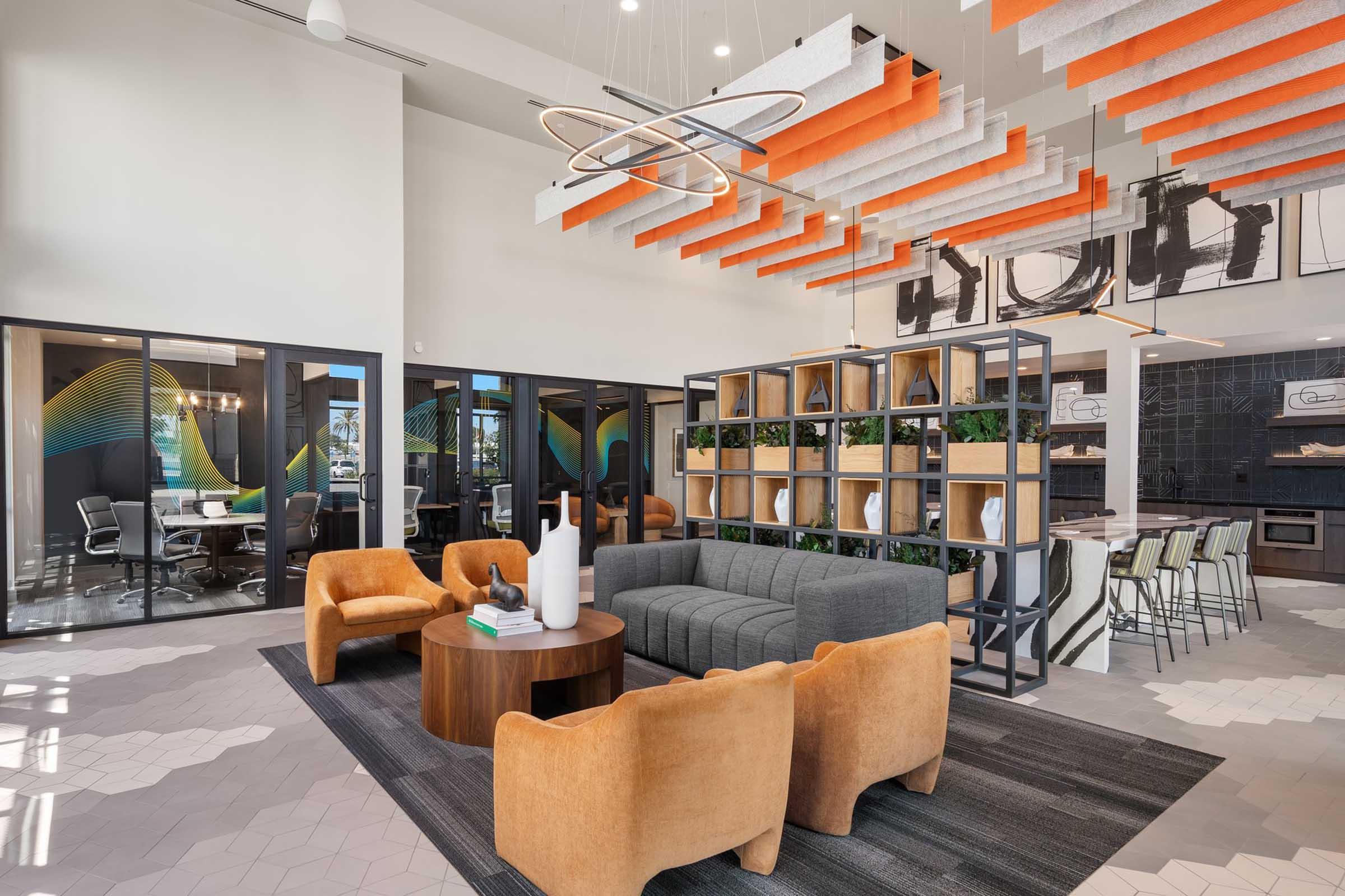 A modern lounge area featuring a gray sofa and orange accent chairs around a circular table. Open spaces reveal glass-walled meeting rooms with contemporary decor, vibrant ceiling elements, and stylish shelving with greenery, creating a welcoming atmosphere in a bright, airy environment.