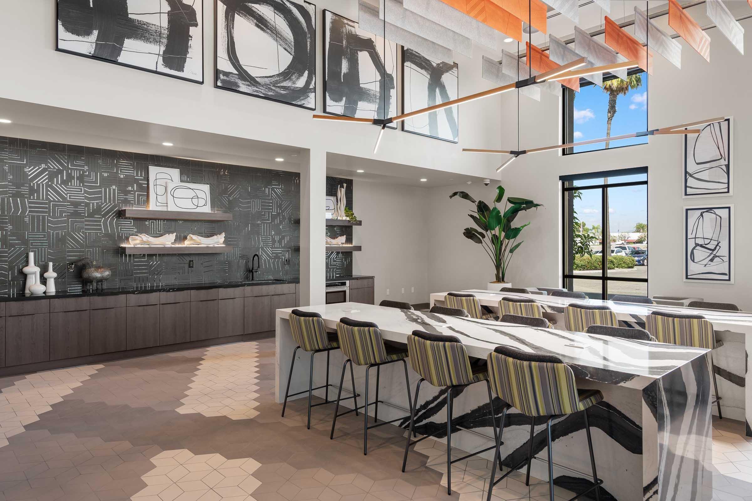Modern kitchen and dining area featuring a large island with black and white marbled countertop, surrounded by striped barstools. The space includes abstract artwork on the walls, a sleek shelving unit with decorative items, and large windows allowing natural light to fill the room.