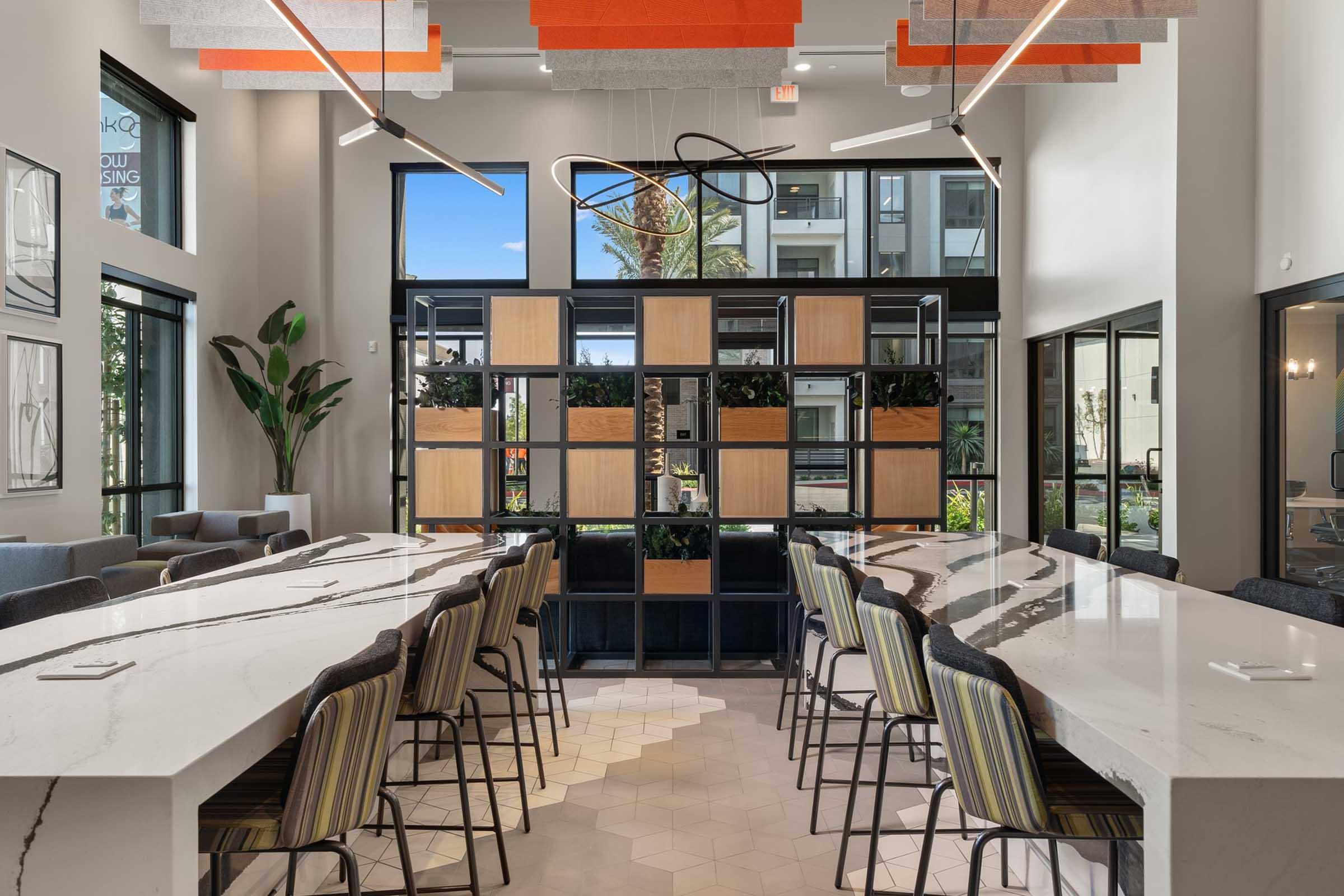 Modern communal space featuring a sleek marble countertop bar with high-backed chairs, a central decorative partition with plants, large windows allowing natural light, and contemporary geometric light fixtures. The design emphasizes openness and a stylish atmosphere.