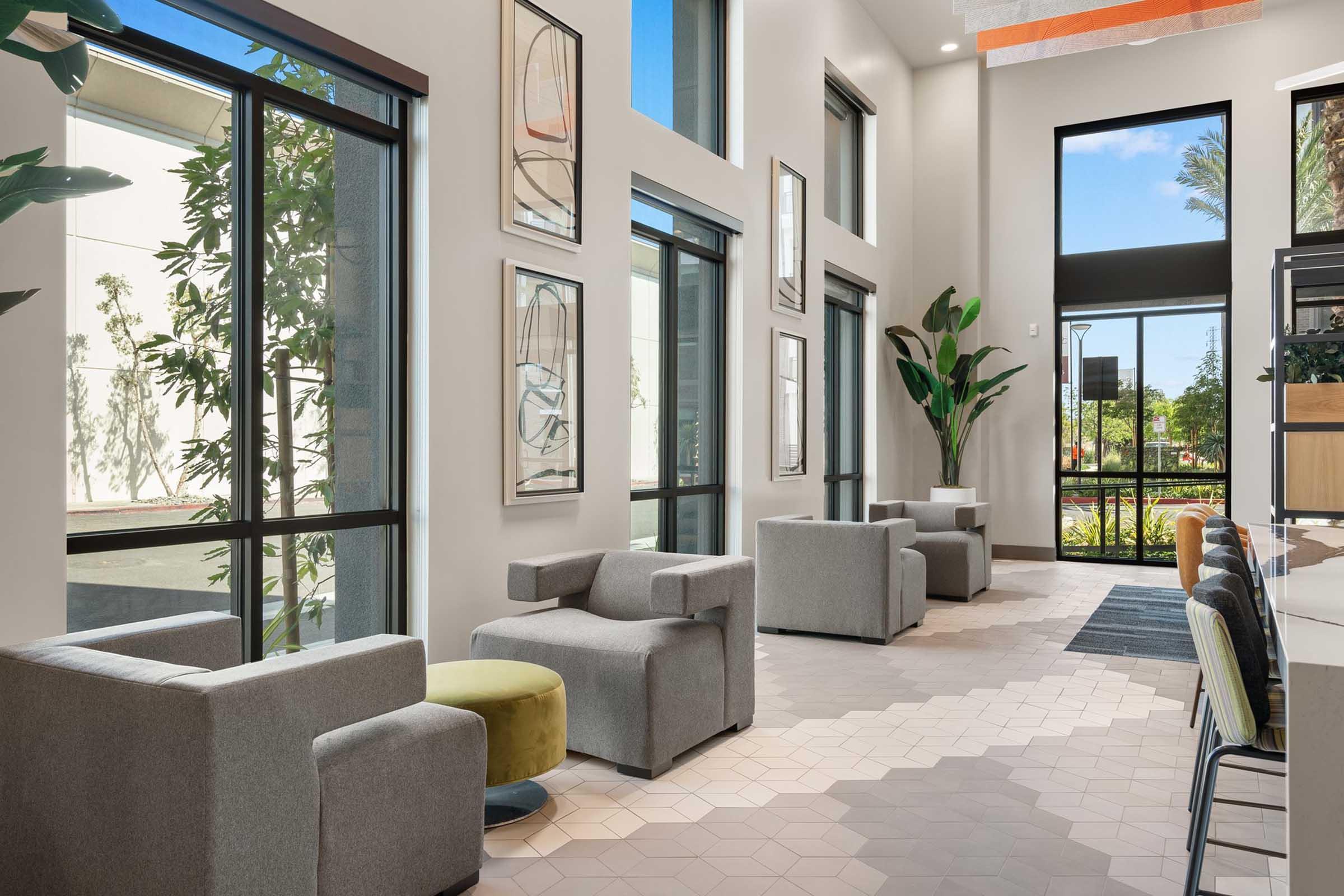Modern interior of a bright lobby featuring large windows, abstract wall art, and comfortable seating arrangements. There are two gray chairs, a colorful ottoman, and a small plant, creating a welcoming and stylish atmosphere. The flooring is a geometric pattern, adding to the contemporary design.