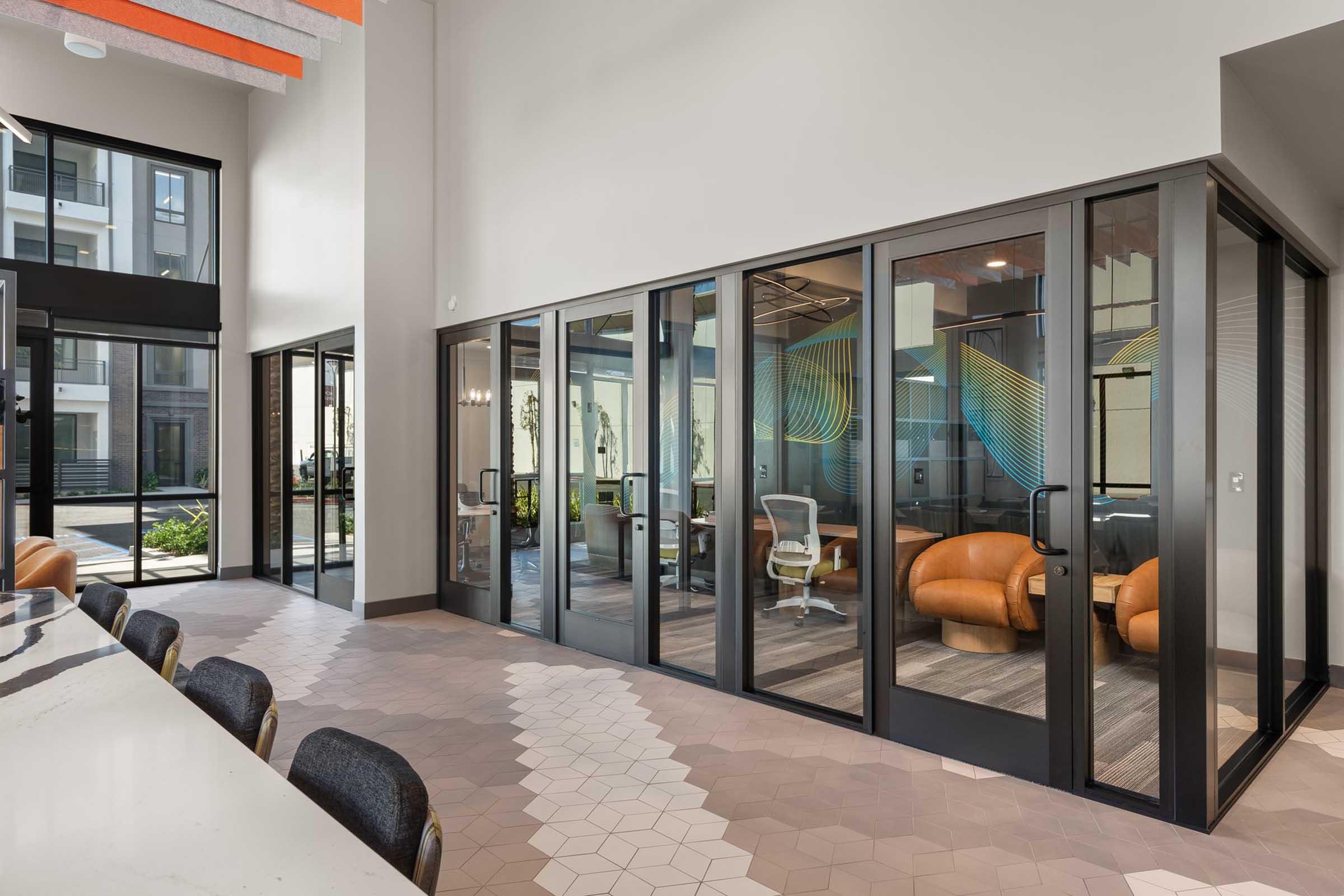 Modern office interior featuring glass-walled meeting rooms with comfortable seating. The common area displays a sleek design with a hexagonal tile floor, contemporary furniture, and large windows allowing natural light. Colorful wall graphics add a vibrant touch to the workspace ambiance.