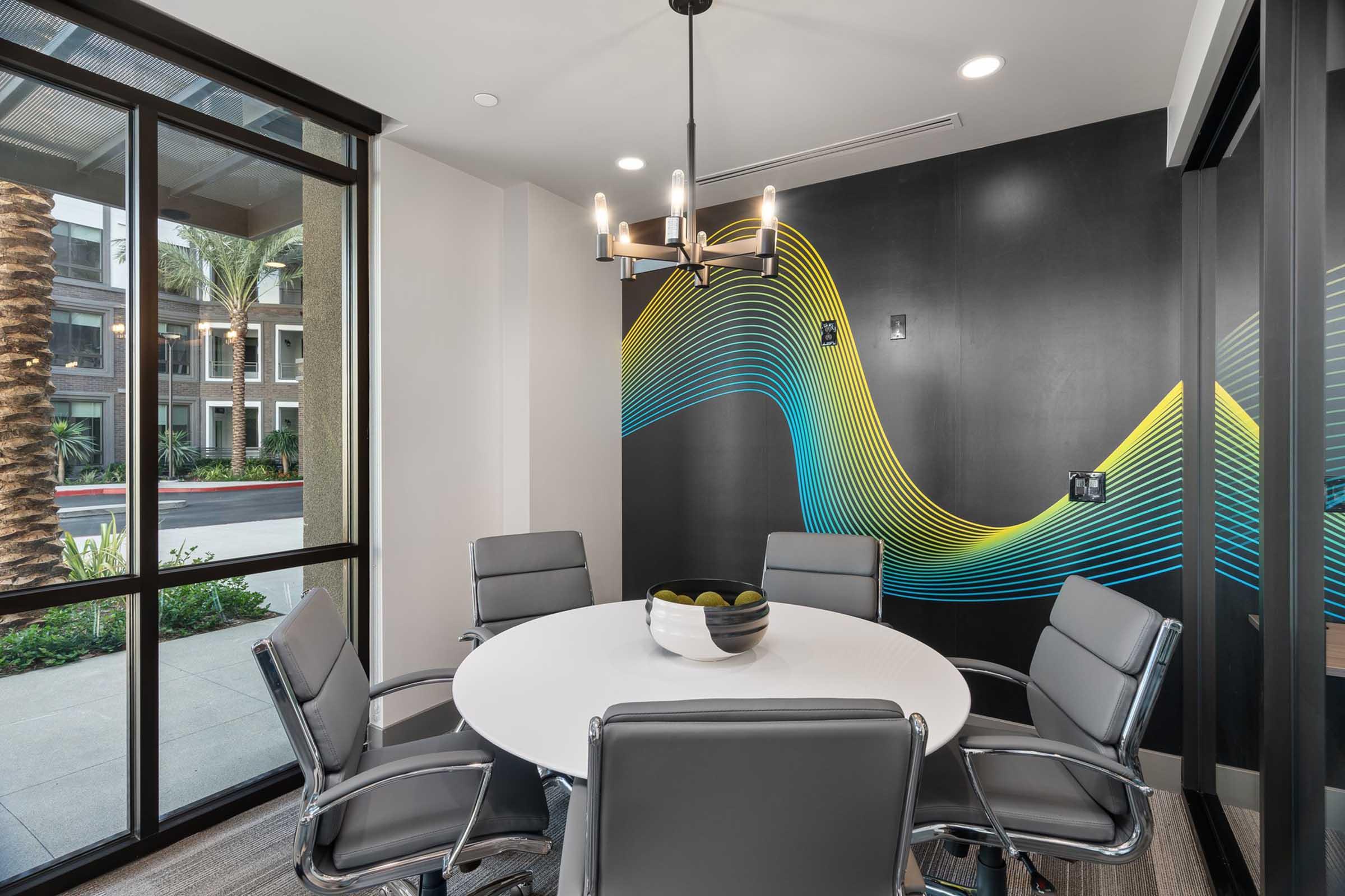 A modern conference room featuring a circular white table surrounded by gray leather chairs. The walls are mostly glass, allowing natural light, and a vibrant wave mural in blue and yellow decorates one wall. A stylish chandelier hangs above the table, and a bowl of green apples sits in the center.