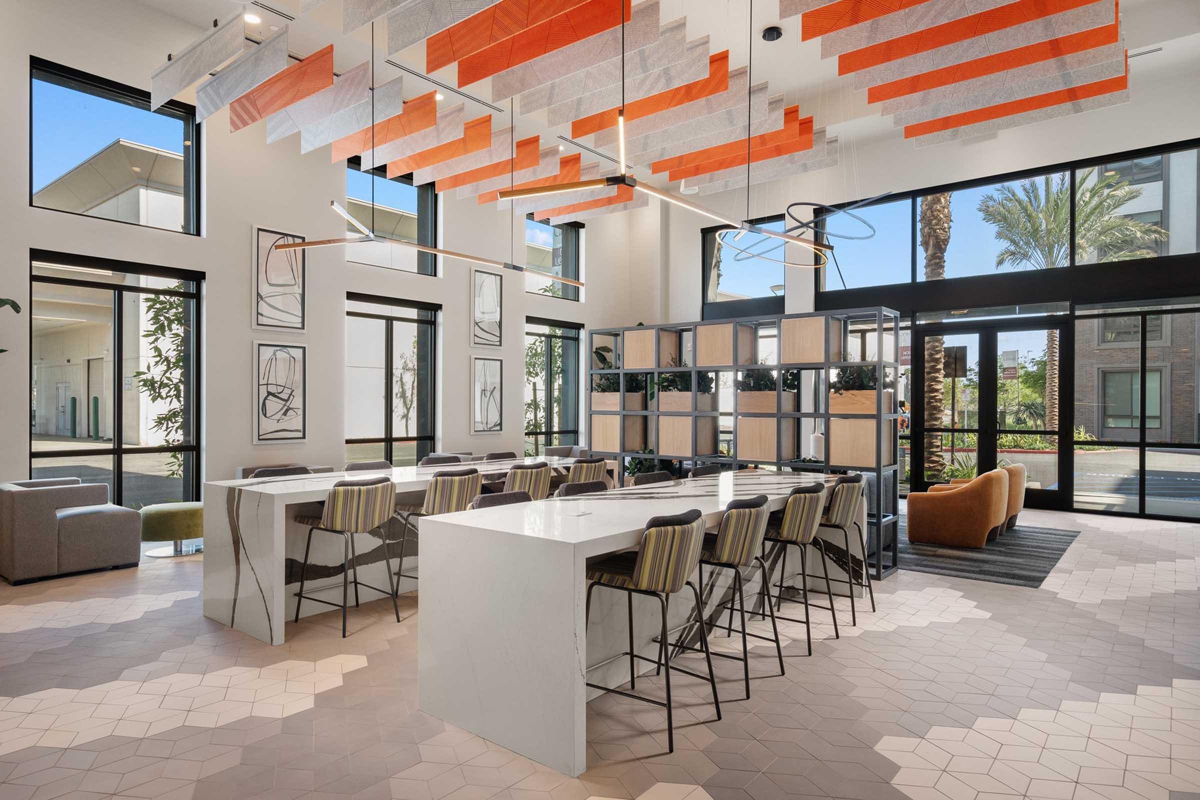 A modern, spacious communal area featuring a stylish white marble table surrounded by bar stools, colorful geometric ceiling decorations in orange and gray, and large windows letting in natural light. There are plants and contemporary furniture, creating an inviting atmosphere.