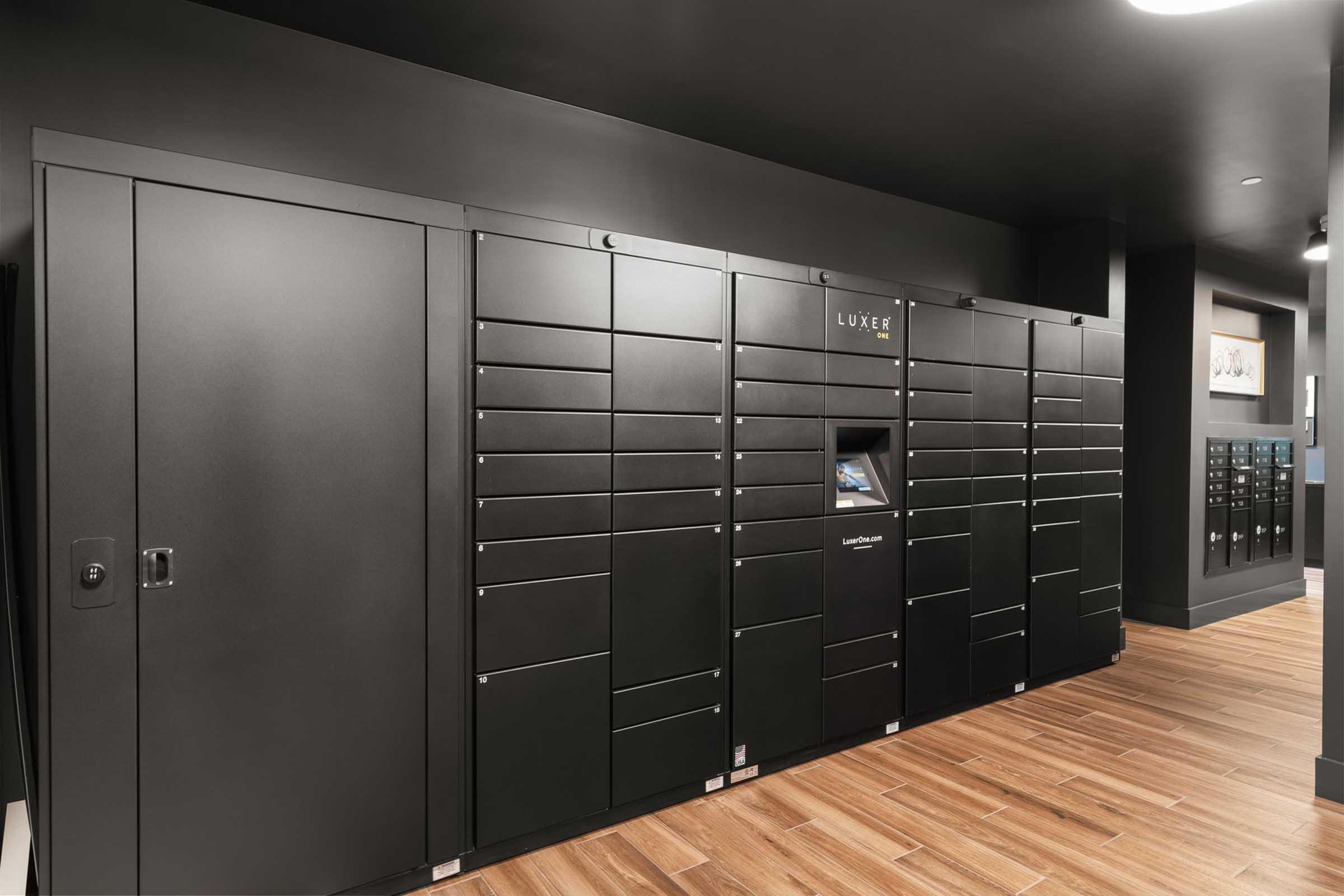 A sleek, modern package locker system against a dark wall, featuring multiple compartments for secure deliveries. There is a central touchscreen for user interaction and a stylish wooden floor in front. The overall ambiance is contemporary and functional, designed for convenience in a residential or commercial setting.