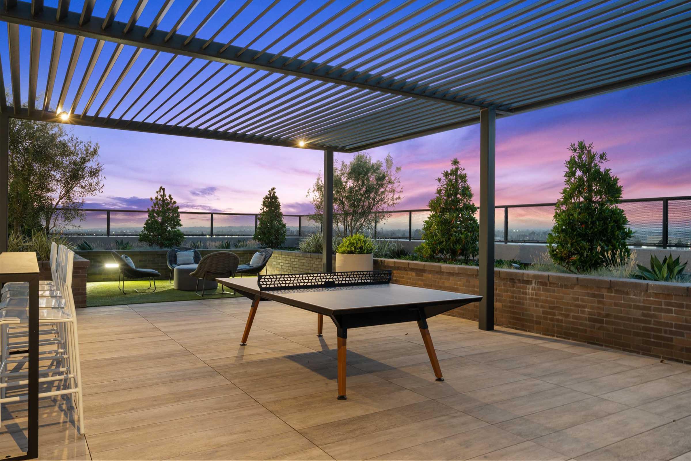 A modern rooftop terrace featuring a ping pong table, seating area with stylish chairs, and planters with greenery. The sky displays a beautiful gradient of purple and blue during sunset, creating a serene and inviting atmosphere.