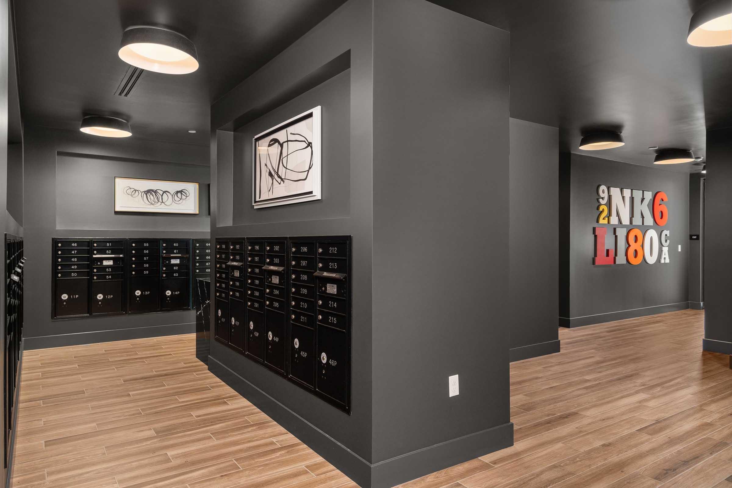 Interior view of a modern mailroom featuring several black mailboxes on the left and a polished wood floor. The walls are painted dark gray, and there's a piece of abstract art framed on the wall. Bright overhead lighting enhances the contemporary design. A colorful decorative sign is displayed on the right wall.