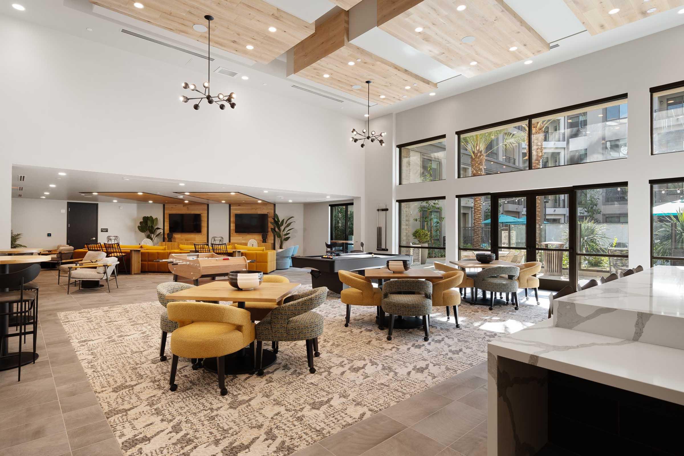 A spacious, modern lounge area featuring yellow and gray seating, a large area rug, and multiple windows allowing natural light. The room includes tables and chairs arranged for socializing, alongside decorative plants. The ceiling has exposed wooden beams, enhancing the contemporary design.