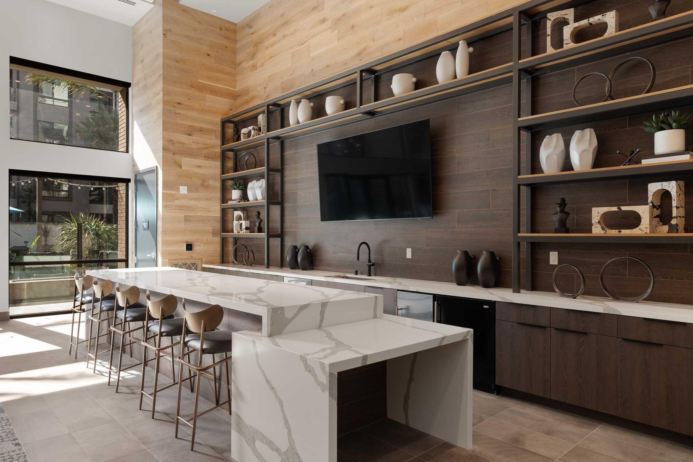 Modern interior of a spacious living area featuring a large marble-topped kitchen island with high stools, a wooden accent wall, and open shelves displaying decorative items. A flat-screen TV is mounted on the wall, and large windows allow natural light to fill the space.
