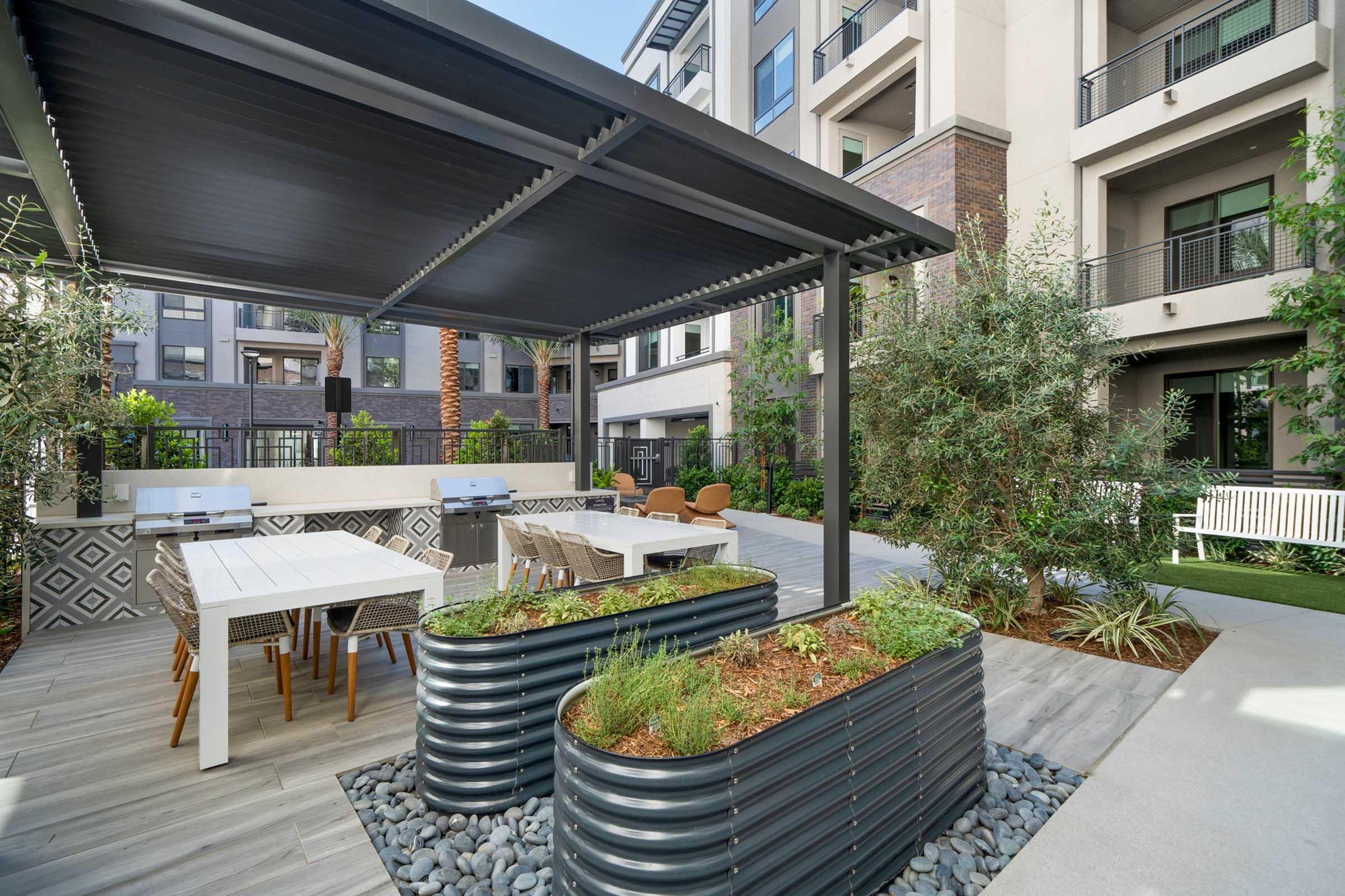 A modern outdoor space featuring a covered patio with a dining table and chairs, barbecues, and raised garden beds filled with plants. The area is surrounded by greenery and contemporary apartment buildings, creating a stylish and inviting atmosphere for relaxation and social gatherings.