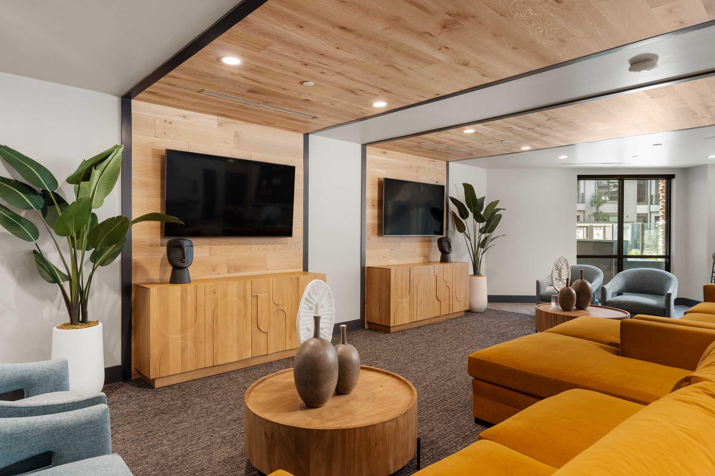 A modern living room featuring two large flat-screen TVs mounted on the wall, a wooden media console, and a circular coffee table with decorative vases. The space includes a bright yellow sectional sofa, light blue armchairs, and indoor plants, with wooden paneling and carpeted flooring.