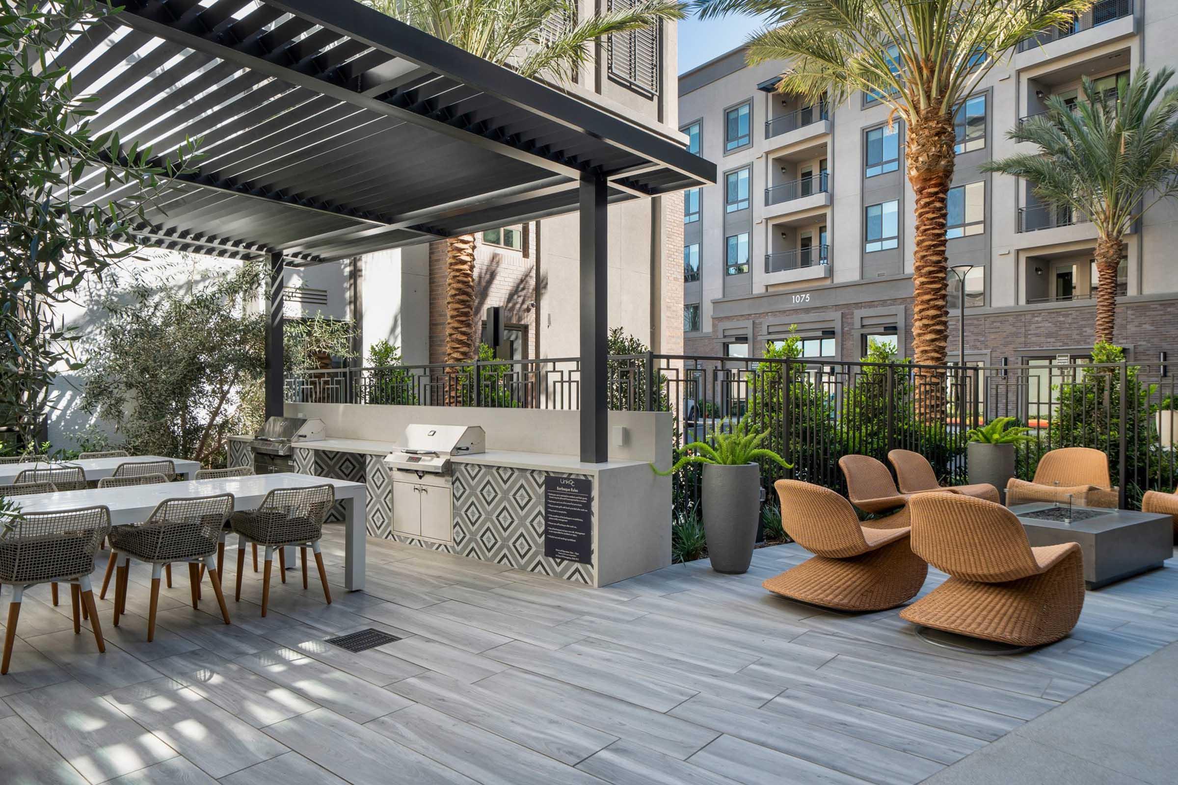 A modern outdoor lounge area featuring stylish seating, a dining table, and an outdoor grill. Surrounded by palm trees and landscaped greenery, the space is designed for relaxation and social gatherings, with contemporary architecture in the background.