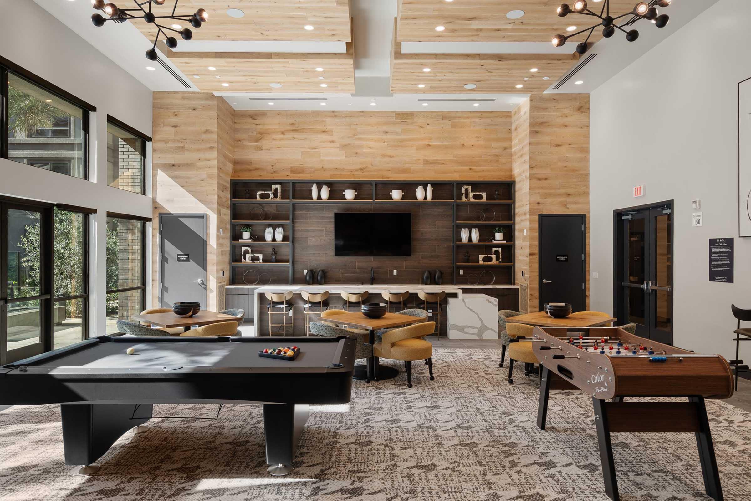 A modern communal game room featuring a billiards table, foosball table, and a variety of seating. The space is well-lit with large windows, wooden accents on the walls, and a flat-screen TV mounted on a dark wooden panel. Stylish decor elements are displayed on shelves, creating an inviting atmosphere.