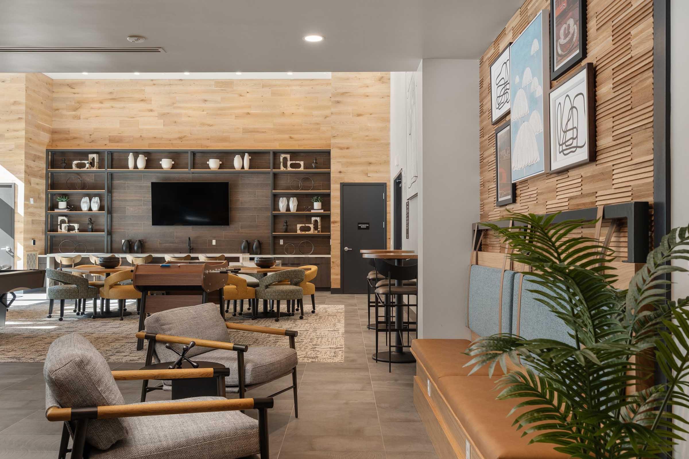 A modern lounge area featuring comfortable seating with armchairs and a long wooden bench. The walls showcase decorative art prints, while shelves hold stylish decor. A large television is mounted on the wall, and the room is illuminated with natural light, creating a welcoming atmosphere.