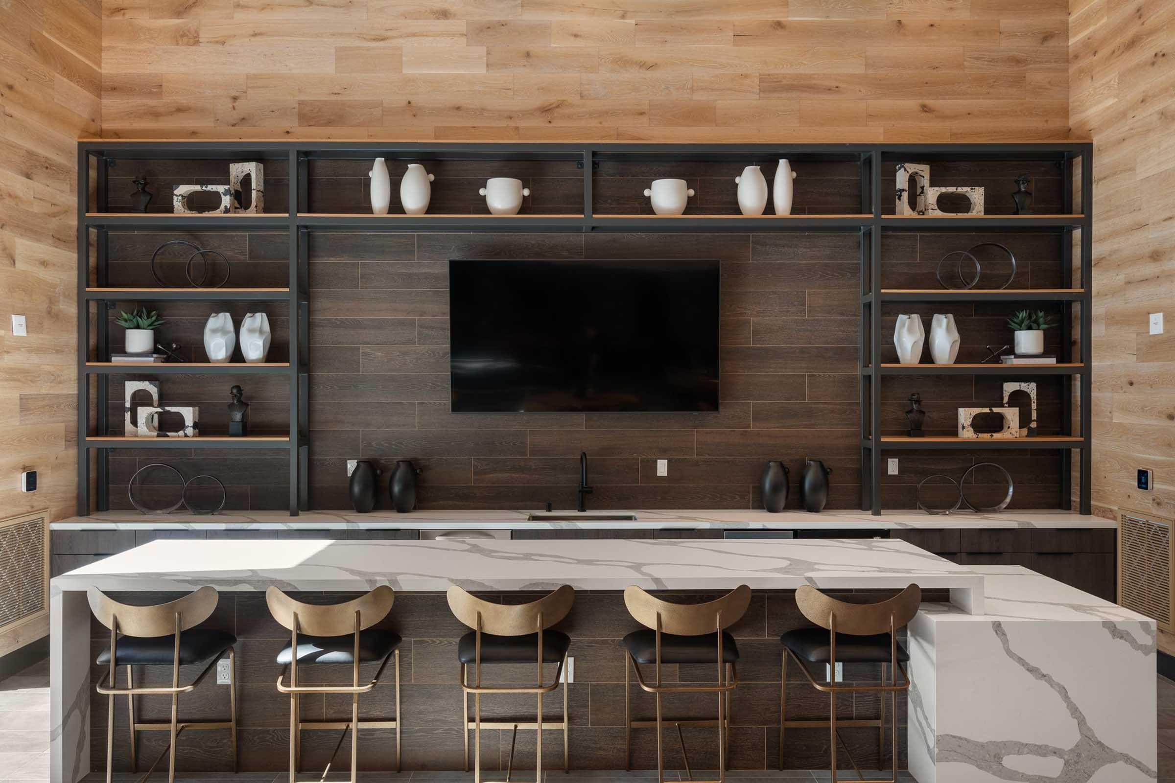A modern kitchen area featuring a wooden accent wall, open shelving with decorative items, and a large flat-screen TV. A marble countertop island with high stools is positioned in front, creating a stylish and functional space.