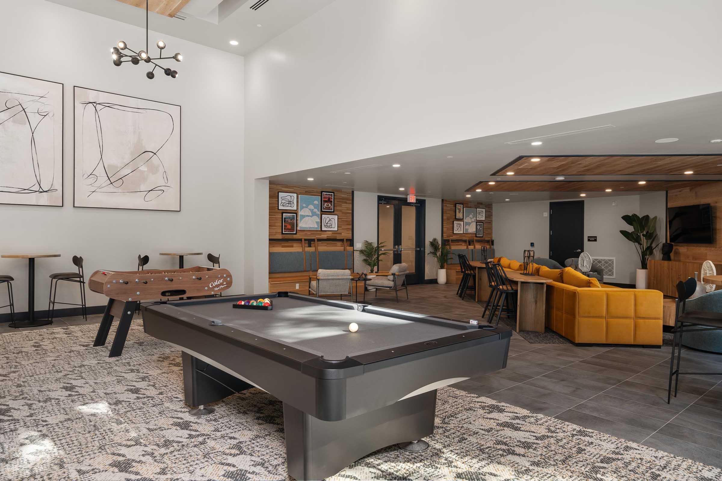 A modern lounge area featuring a billiard table in the foreground, surrounded by stylish seating and tables. The space is bright with large windows and contemporary artwork on the walls, creating a welcoming atmosphere for socializing and relaxation.