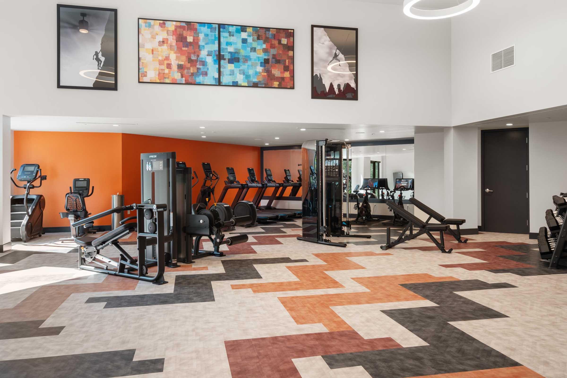 Modern gym interior featuring various exercise equipment, including cardio machines and weightlifting benches. The space has colorful artwork on the walls, an orange accent wall, and a patterned floor in shades of gray, orange, and black, creating a vibrant workout environment.