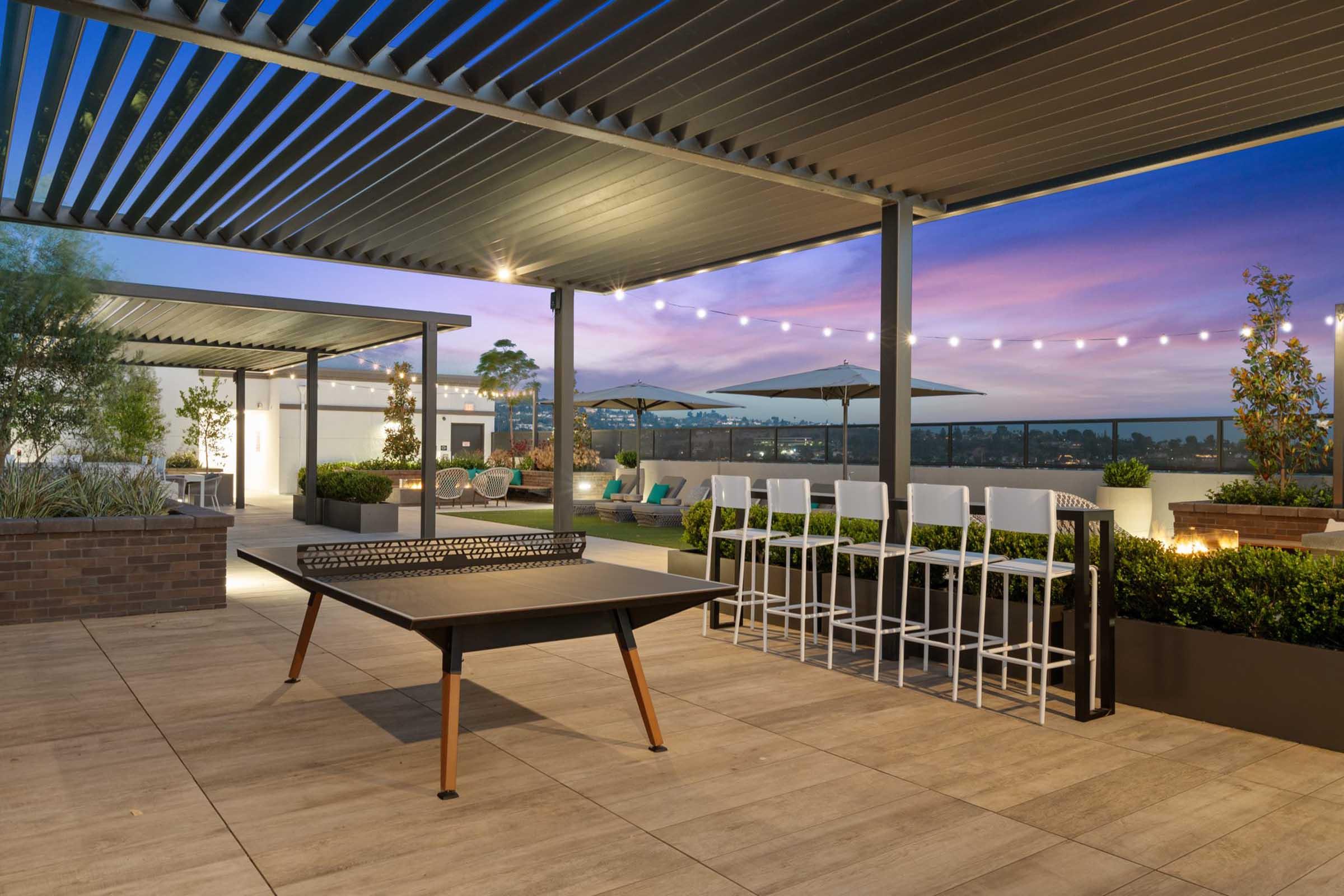 A modern rooftop terrace featuring a ping pong table, bar stools, lush planters, and ambient string lights. The scene is illuminated by the soft glow of sunset, with a clear sky and a relaxing atmosphere, ideal for social gatherings.
