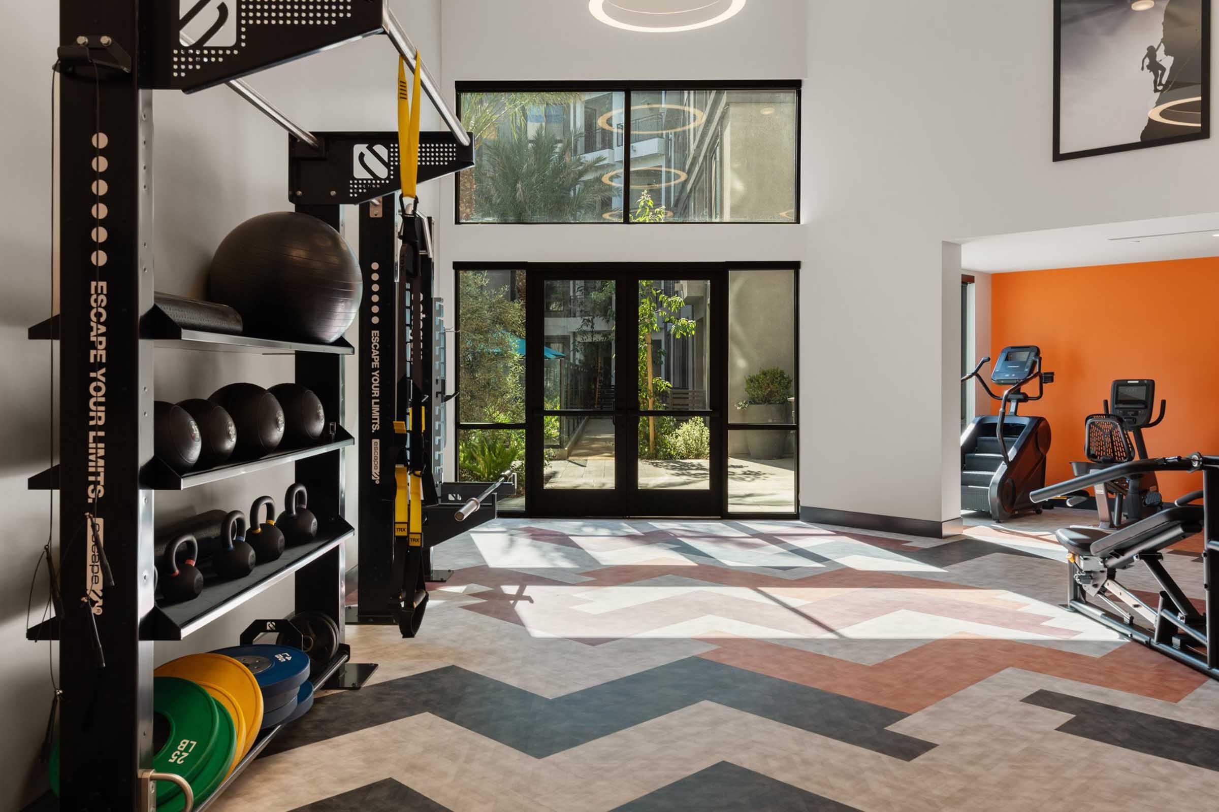 A modern gym interior featuring exercise equipment such as kettlebells, medicine balls, and resistance bands on racks. Large windows provide natural light, and there are fitness machines in the background. The floor has a chevron pattern, and an inspirational quote is visible on a wall.
