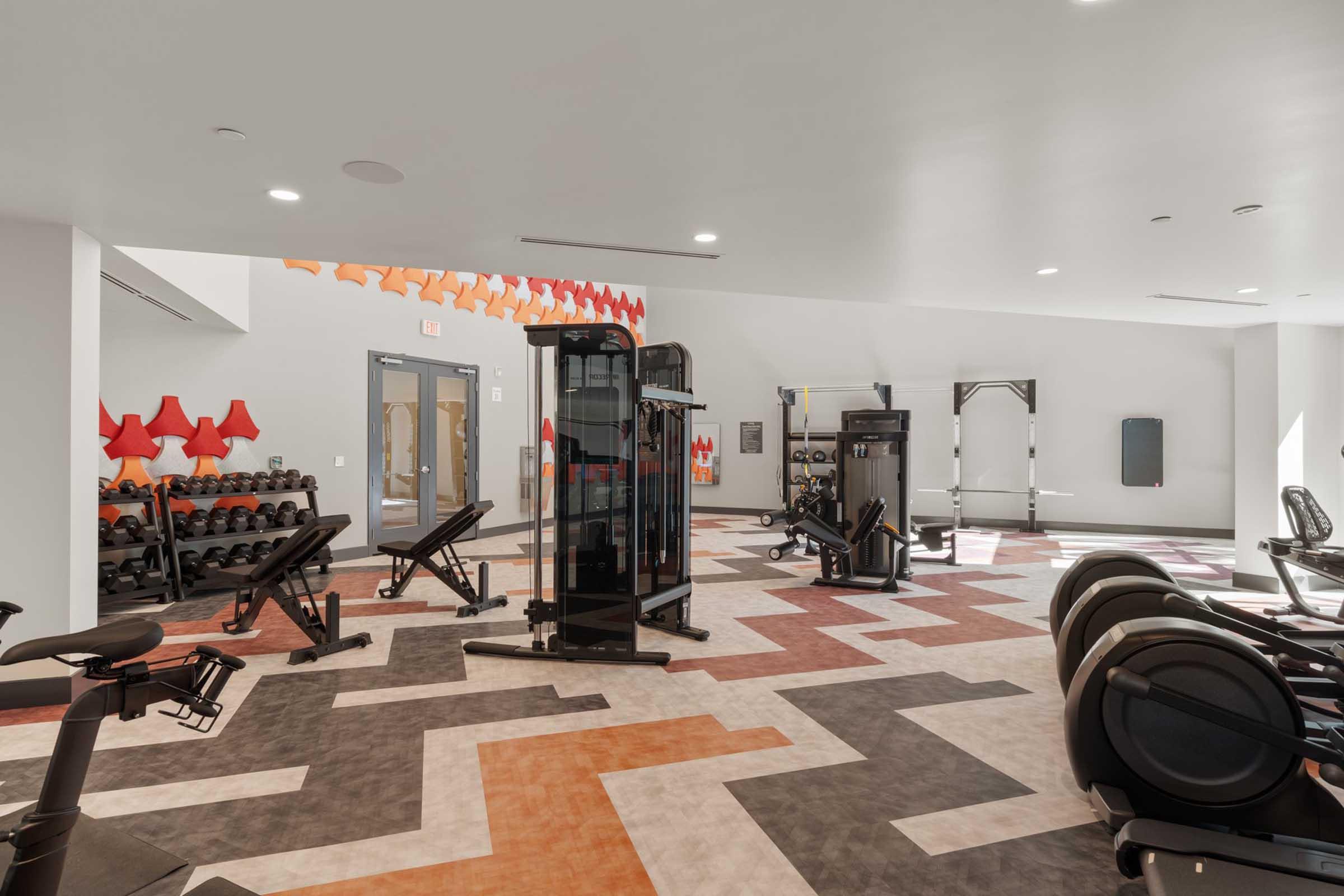 A spacious gym featuring various fitness equipment, including weight benches, dumbbells, and cardio machines. The interior design includes colorful geometric patterns on the floor and decorative elements on the walls, creating a modern and inviting workout environment.
