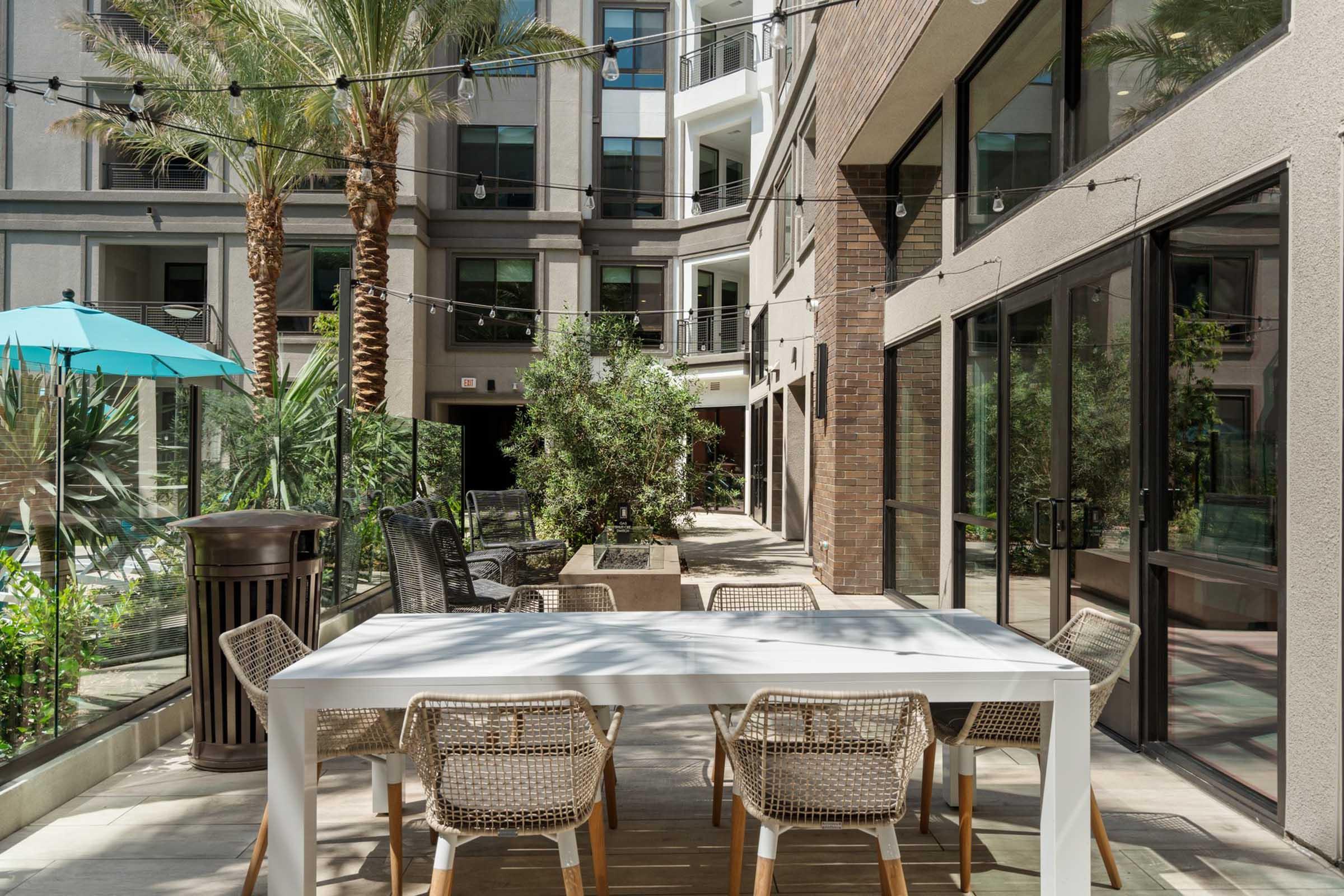 A modern outdoor courtyard featuring a large white dining table surrounded by wicker chairs, palm trees, and decorative plants. An umbrella in turquoise color provides shade, and string lights are strung overhead, creating a relaxed, inviting atmosphere. The surrounding building has large windows that enhance the view.