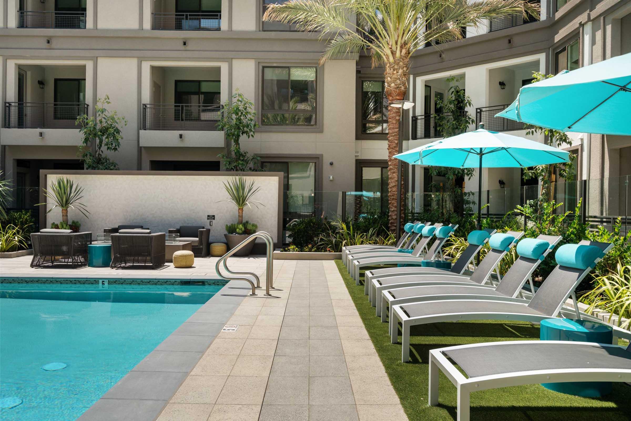 A serene pool area featuring a sparkling blue swimming pool surrounded by modern lounge chairs and turquoise umbrellas. Lush greenery and palm trees enhance the tropical vibe, complemented by stylish outdoor furniture set against a contemporary building backdrop.