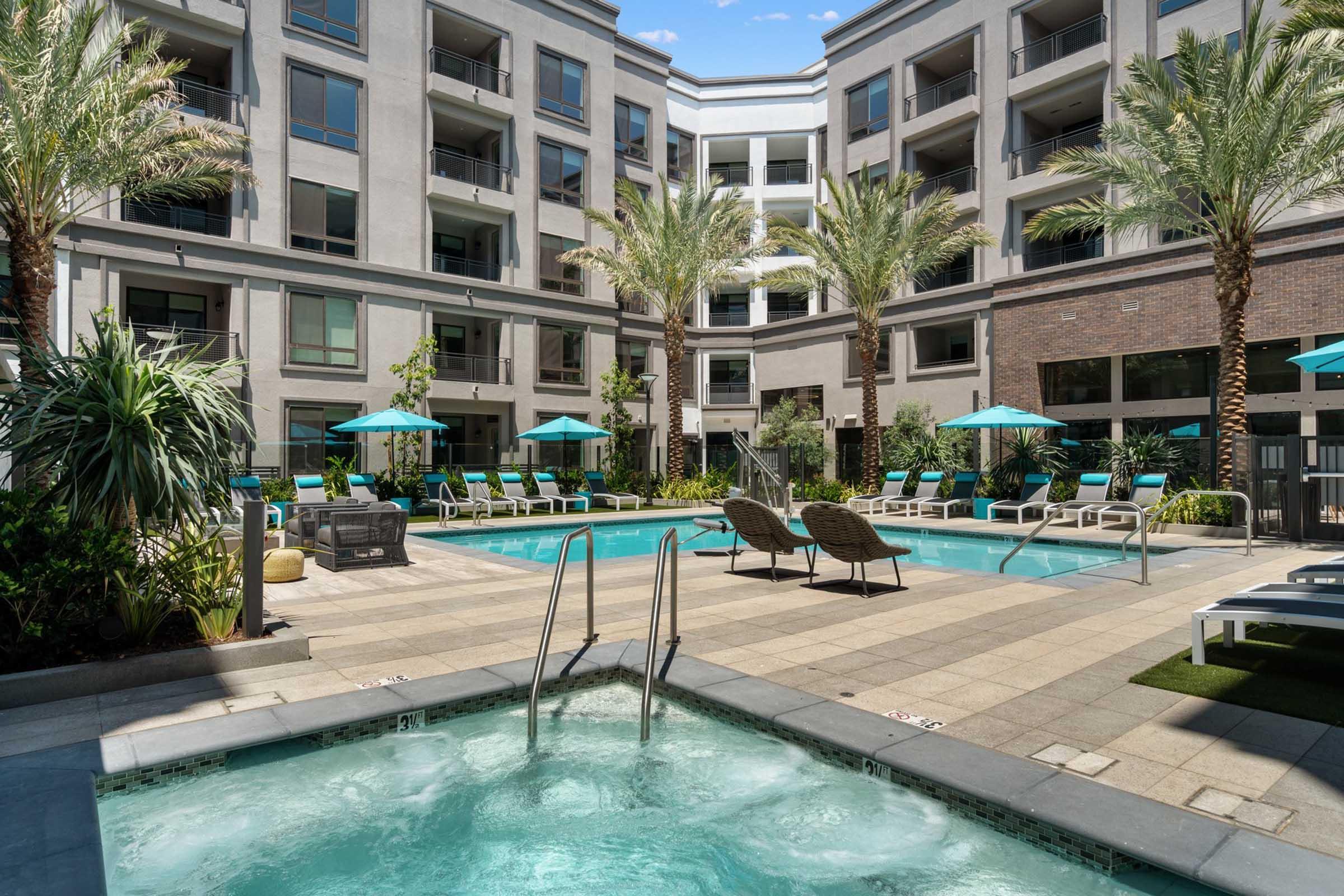 A modern outdoor pool area surrounded by palm trees with lounge chairs and umbrellas. The pool features a spa and is part of a contemporary apartment complex. Sunlight filters through, creating a vibrant and inviting atmosphere for relaxation and recreation.