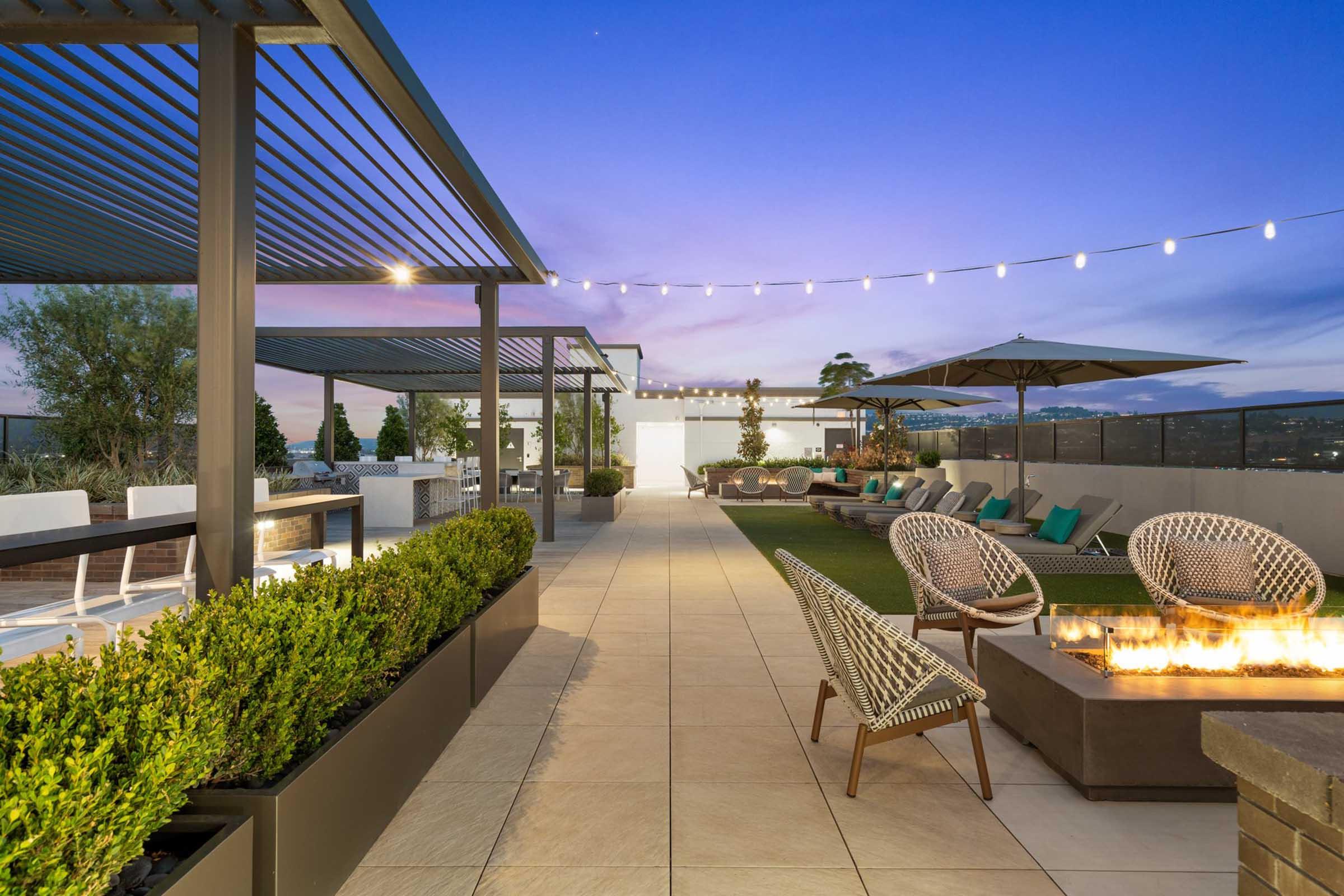 Rooftop terrace at dusk featuring stylish seating, a fire pit, and greenery. Modern design elements include pergolas and string lights, creating a relaxing atmosphere. The space is well-lit and offers a view of the sunset, ideal for social gatherings or quiet evenings outdoors.