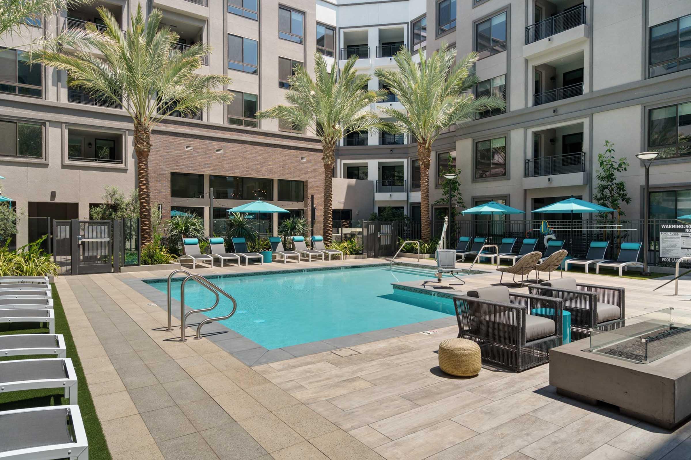 A modern apartment complex pool area featuring clear blue water, surrounded by lounge chairs and umbrellas. Palm trees provide shade, while a few seating areas invite relaxation. The surrounding building has large windows and a contemporary design, enhancing the inviting atmosphere.