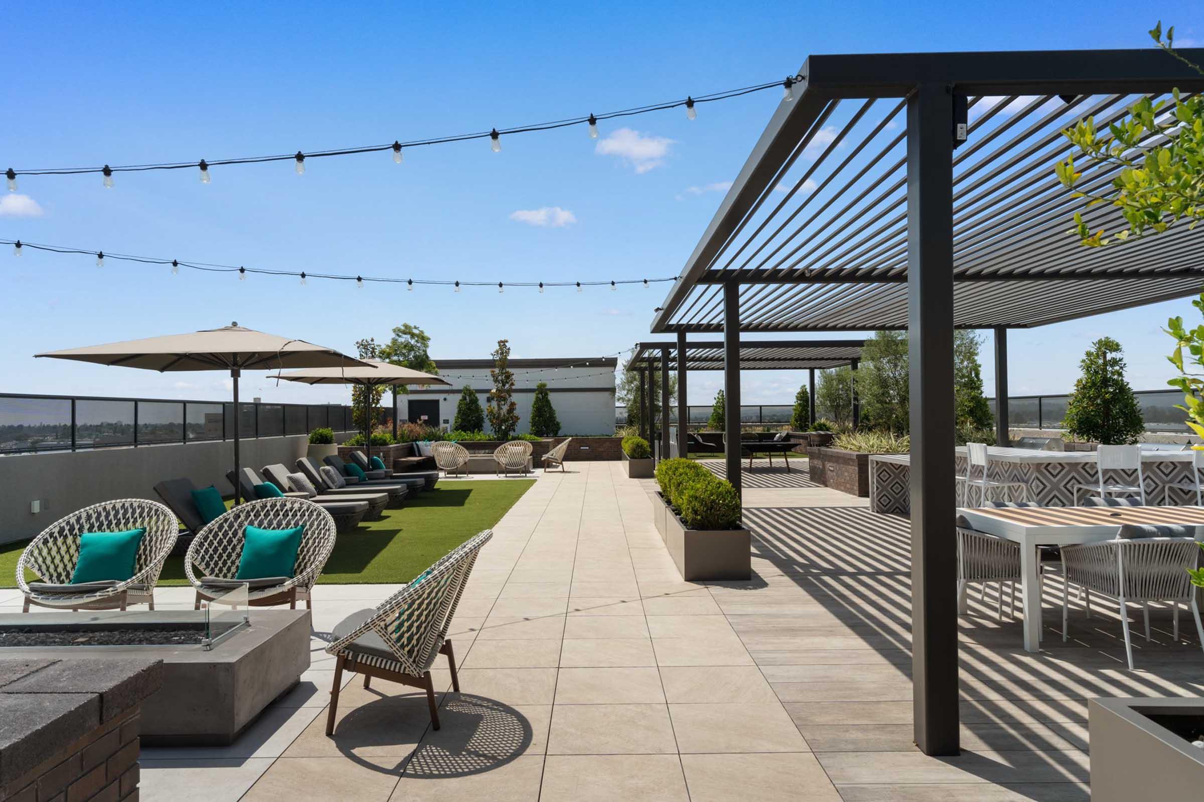 A modern rooftop terrace featuring stylish outdoor furniture, including lounge chairs and dining tables, surrounded by greenery. Soft lighting from string lights adds a cozy atmosphere, and the space is adorned with planters for a touch of nature against a clear blue sky.