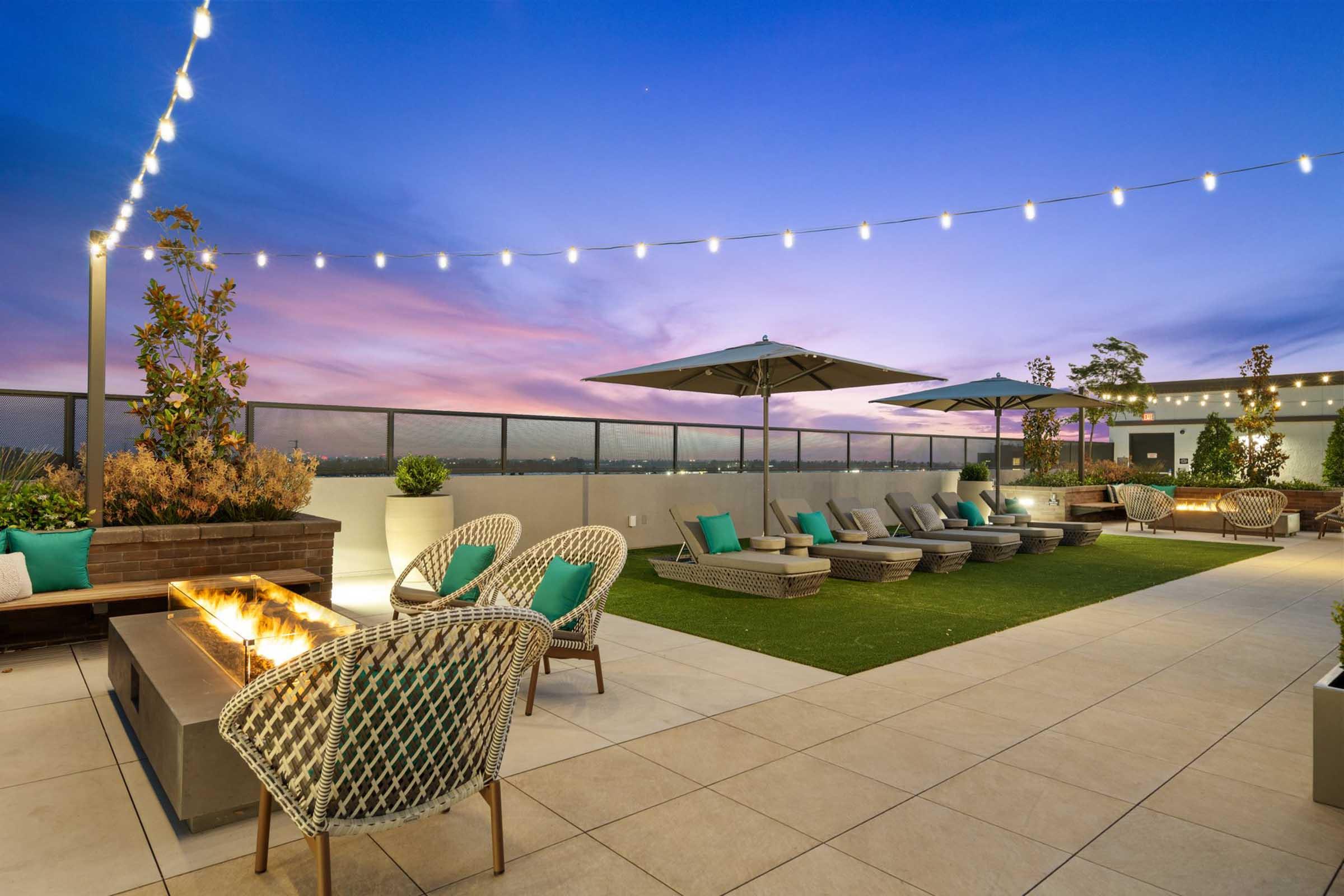 Rooftop lounge area at dusk, featuring comfortable seating with teal cushions, patio umbrellas, and a fire pit. Soft, twinkling string lights illuminate the space, creating a cozy atmosphere against a colorful sunset sky. Lush greenery adds a touch of nature to the urban setting.