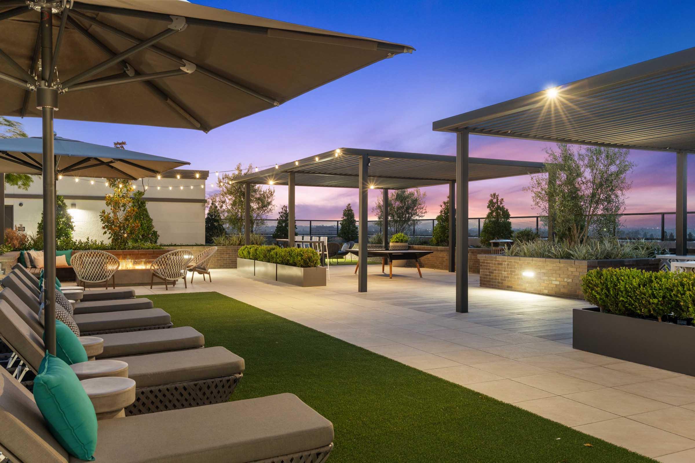 Rooftop terrace featuring lounge chairs with umbrellas, landscaped greenery, and a scenic sunset sky. Modern design elements include planters and seating areas, creating a relaxing atmosphere for socializing or enjoying the view.