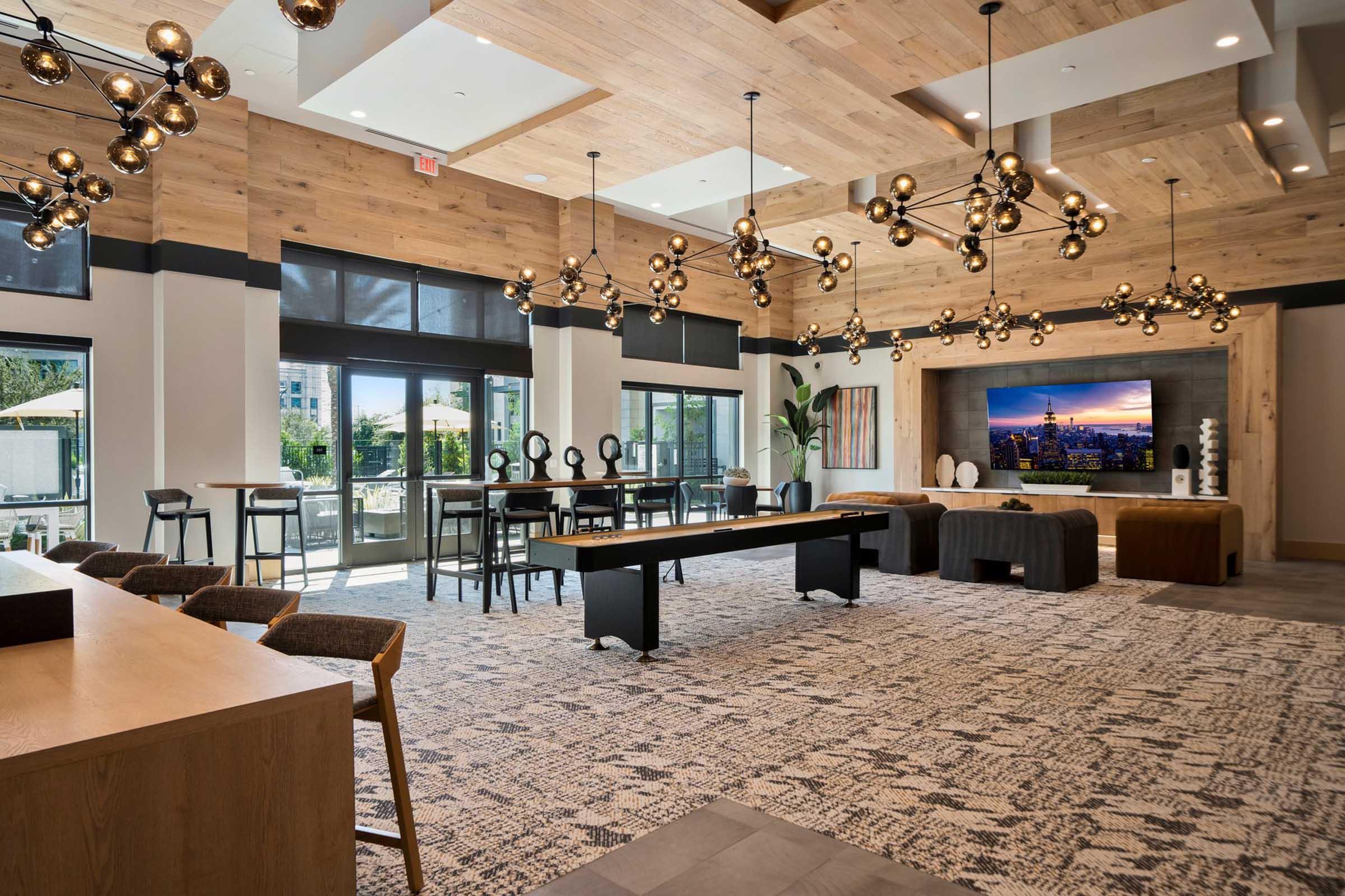 Modern lounge area with wooden beams and decorative light fixtures. Features a pool table, seating areas with stylish chairs and tables, and large windows allowing natural light. A screen displays a scenic view. The space has a contemporary design, combining warm wood tones with sleek furniture.